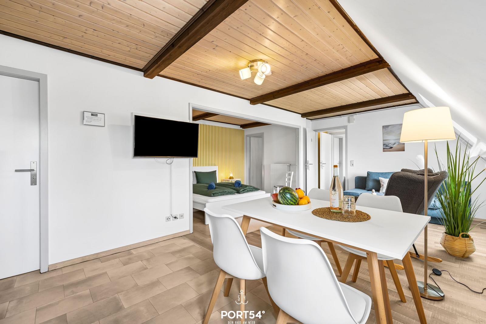 Essbereich mit weißem Tisch und Stühlen, Fernseher und Schlafbereich im Hintergrund.