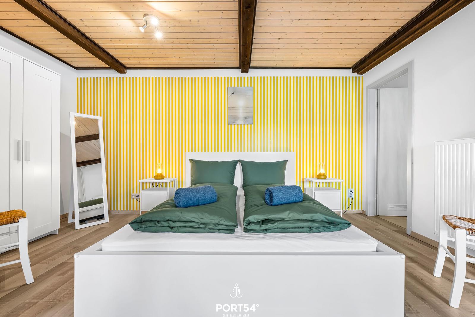 Schlafzimmer mit Doppelbett, gelb gestreifter Wand und Holzboden.