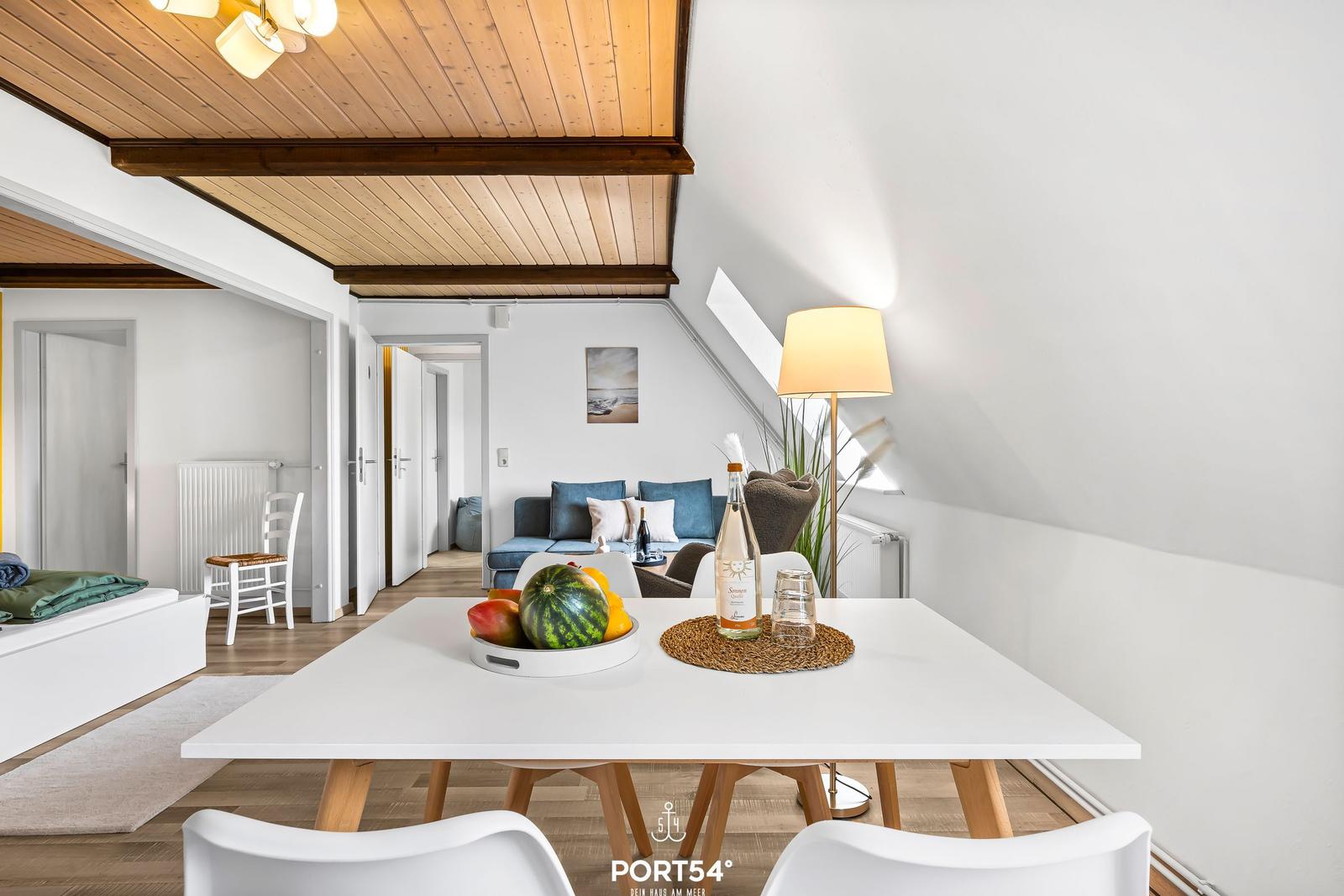 Essbereich mit Tisch und Stühlen. Im Hintergrund Wohnbereich mit Sofa und Fenster.