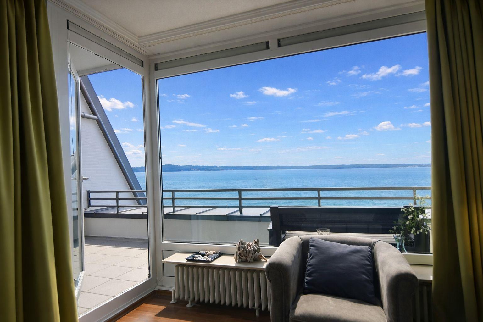 Ein Sessel mit Kissen steht vor großen Fenstern mit Meerblick.