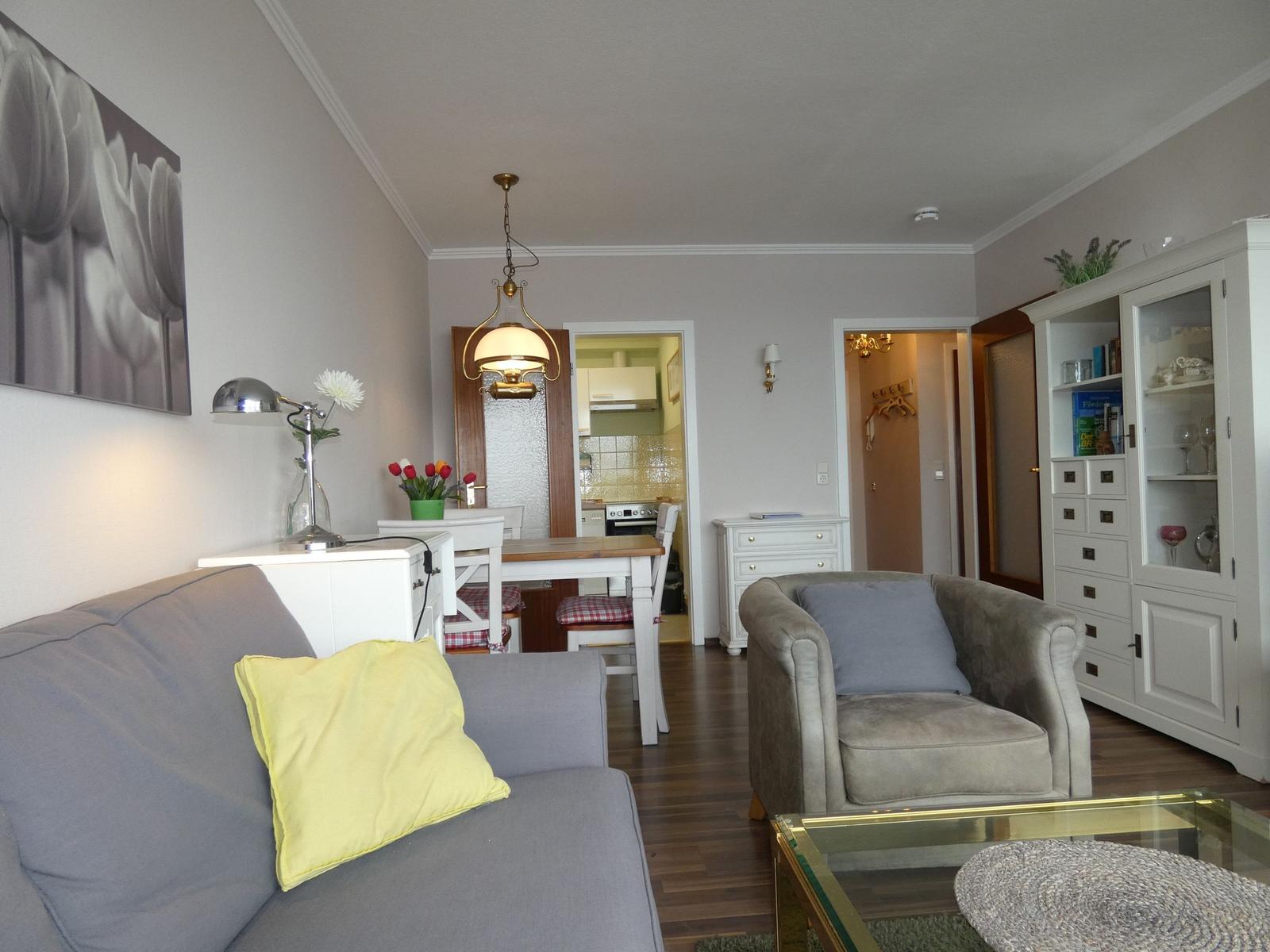 Wohnzimmer mit Sofa, Sessel und Esstisch. Küche und Flur sind sichtbar.