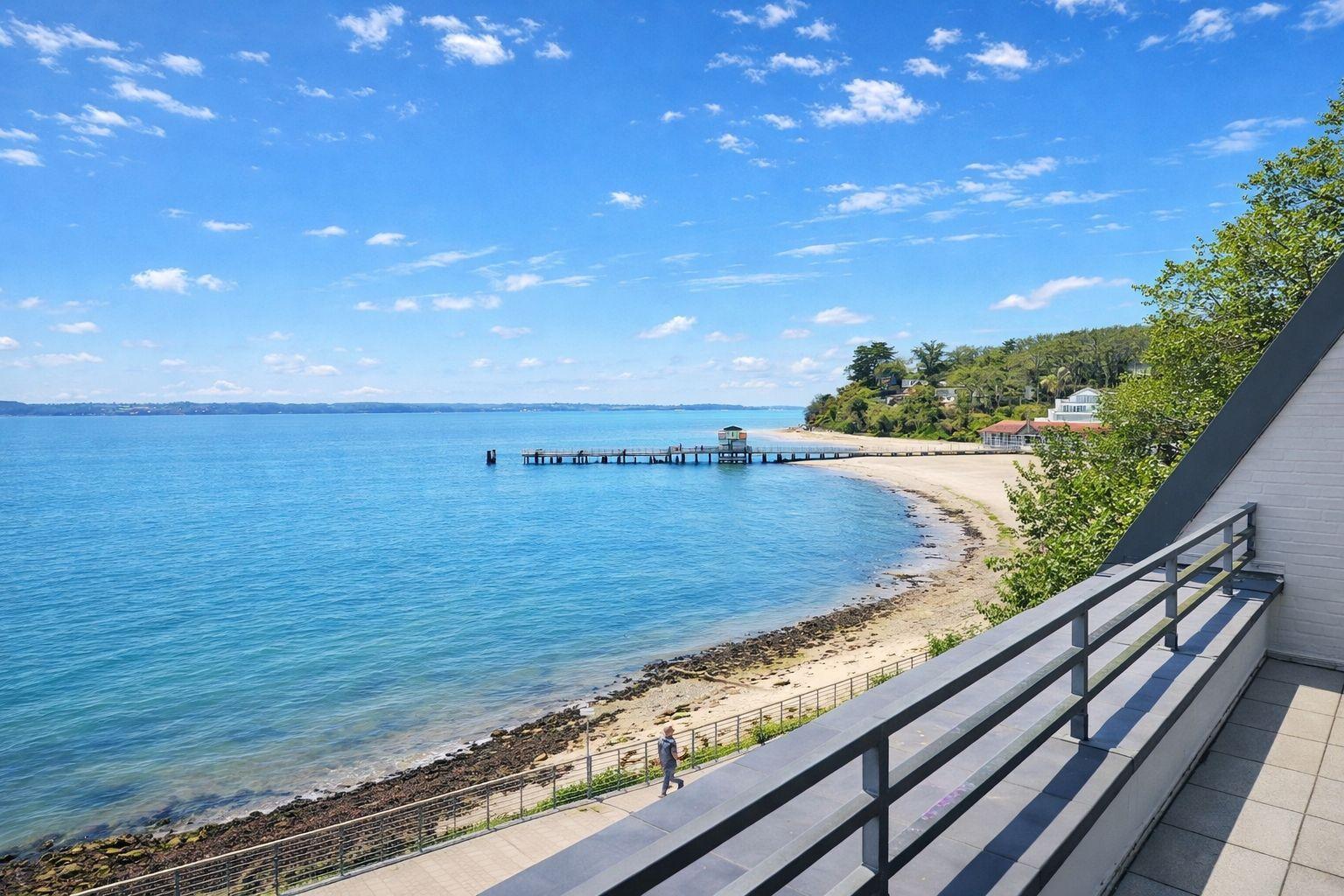 Blick auf Strand, See und Steg unter blauem Himmel mit wenigen Wolken.