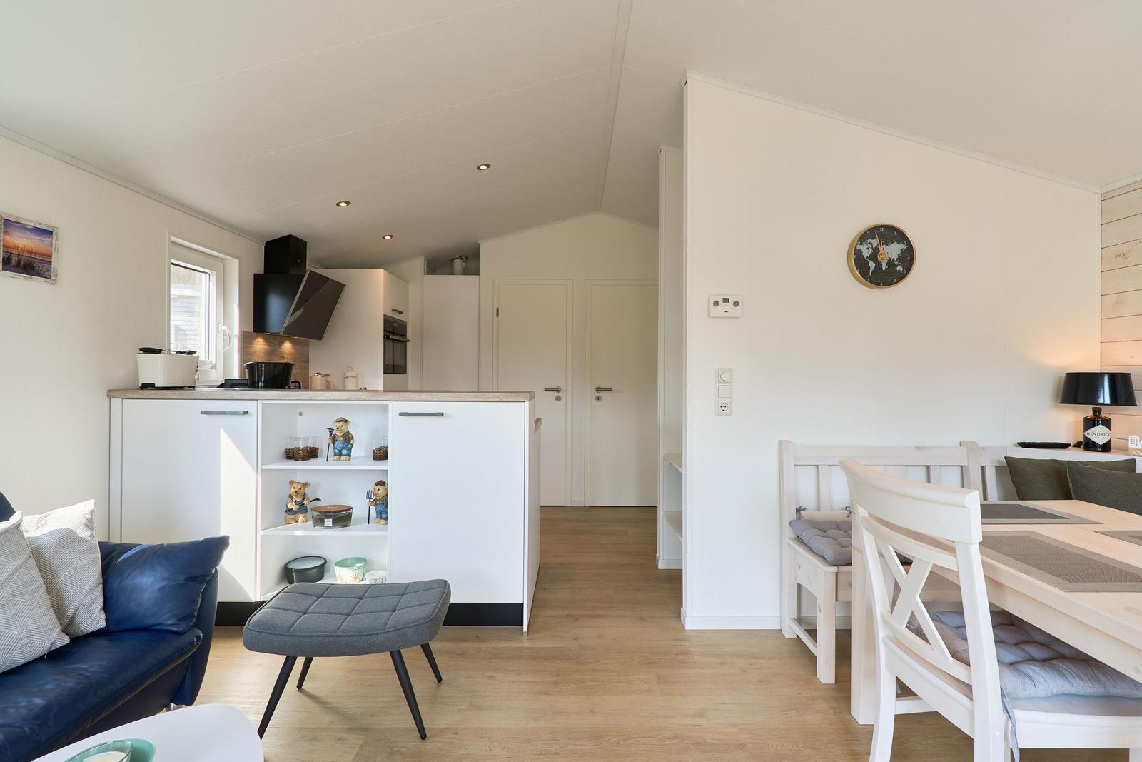 Wohn- und Essbereich mit Küchenzeile, Esstisch und Sofa. Wanduhr und Holzboden sichtbar.