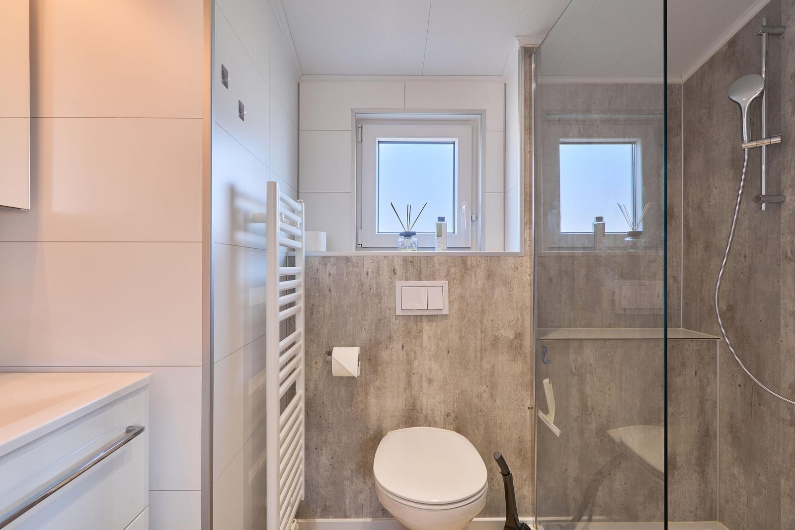 Badezimmer mit Dusche, Toilette und Fenster. Wandverkleidung aus Betonoptik.