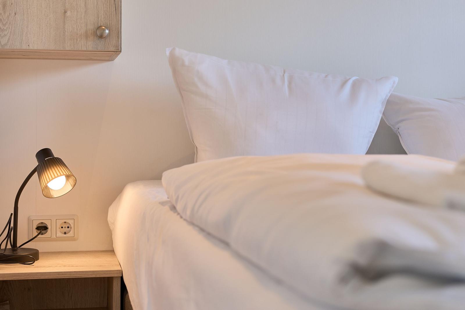 Ein Bett mit weißen Kissen und Bettwäsche neben einem Nachttisch mit Lampe.