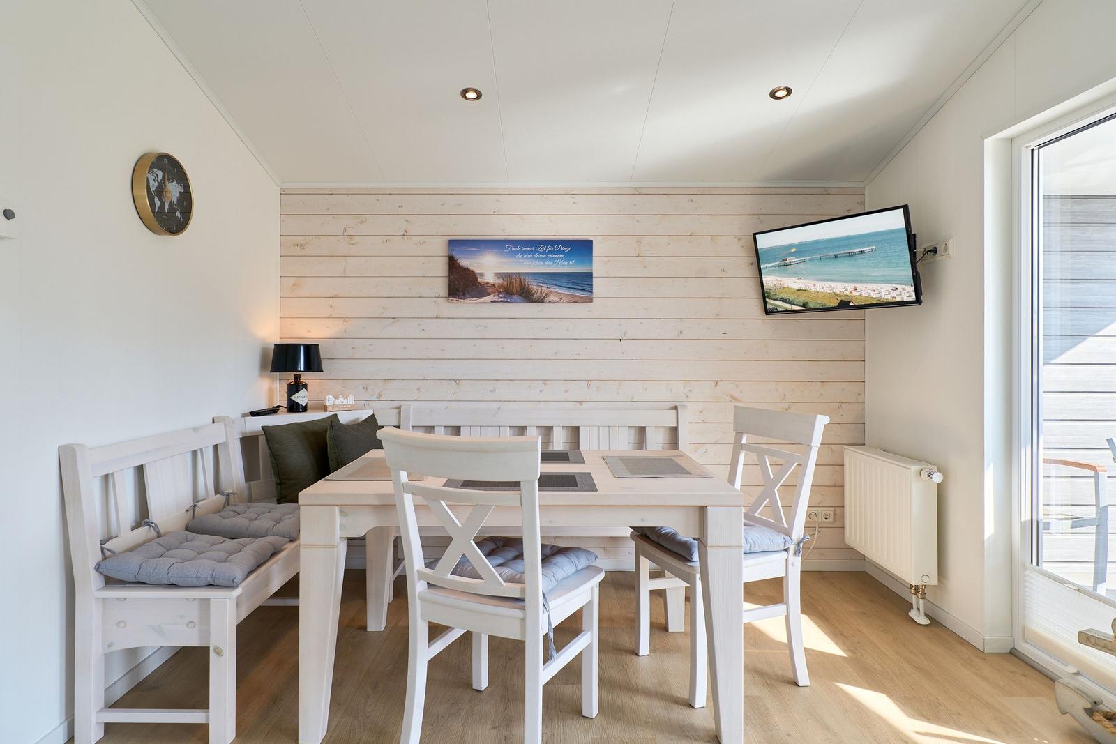 Esszimmer mit weißem Tisch, Stühlen und Sitzbank. Wandbild und TV an der Holzwand.
