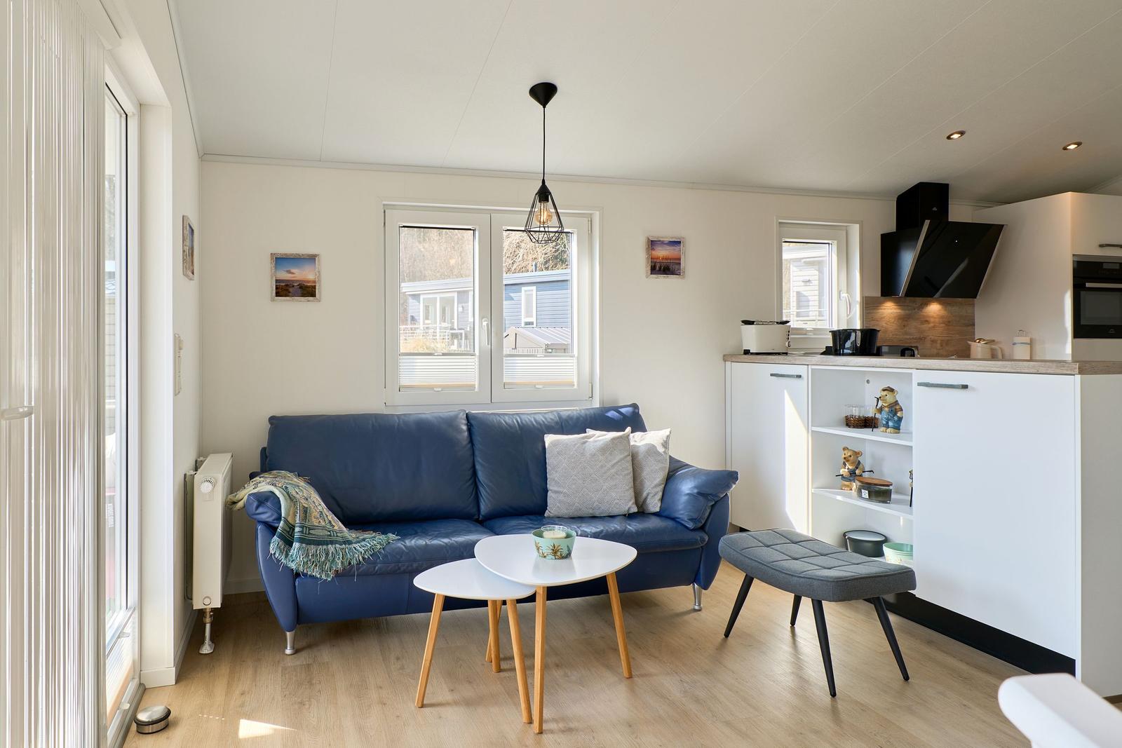 Ein Wohnbereich mit blauem Sofa, weißen Tischen und einer Küchenzeile.