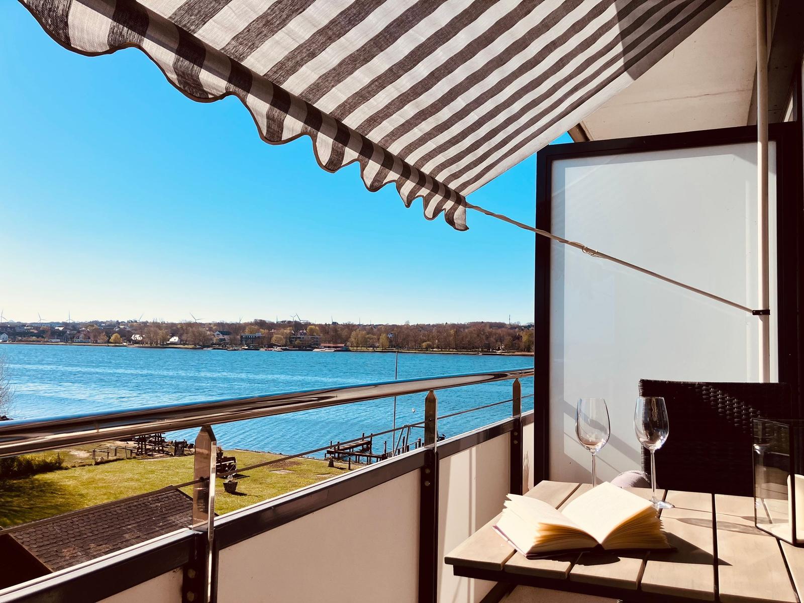 Terrasse mit Blick auf See, Tisch mit zwei Gläsern und offenem Buch unter Streifenmarkise.