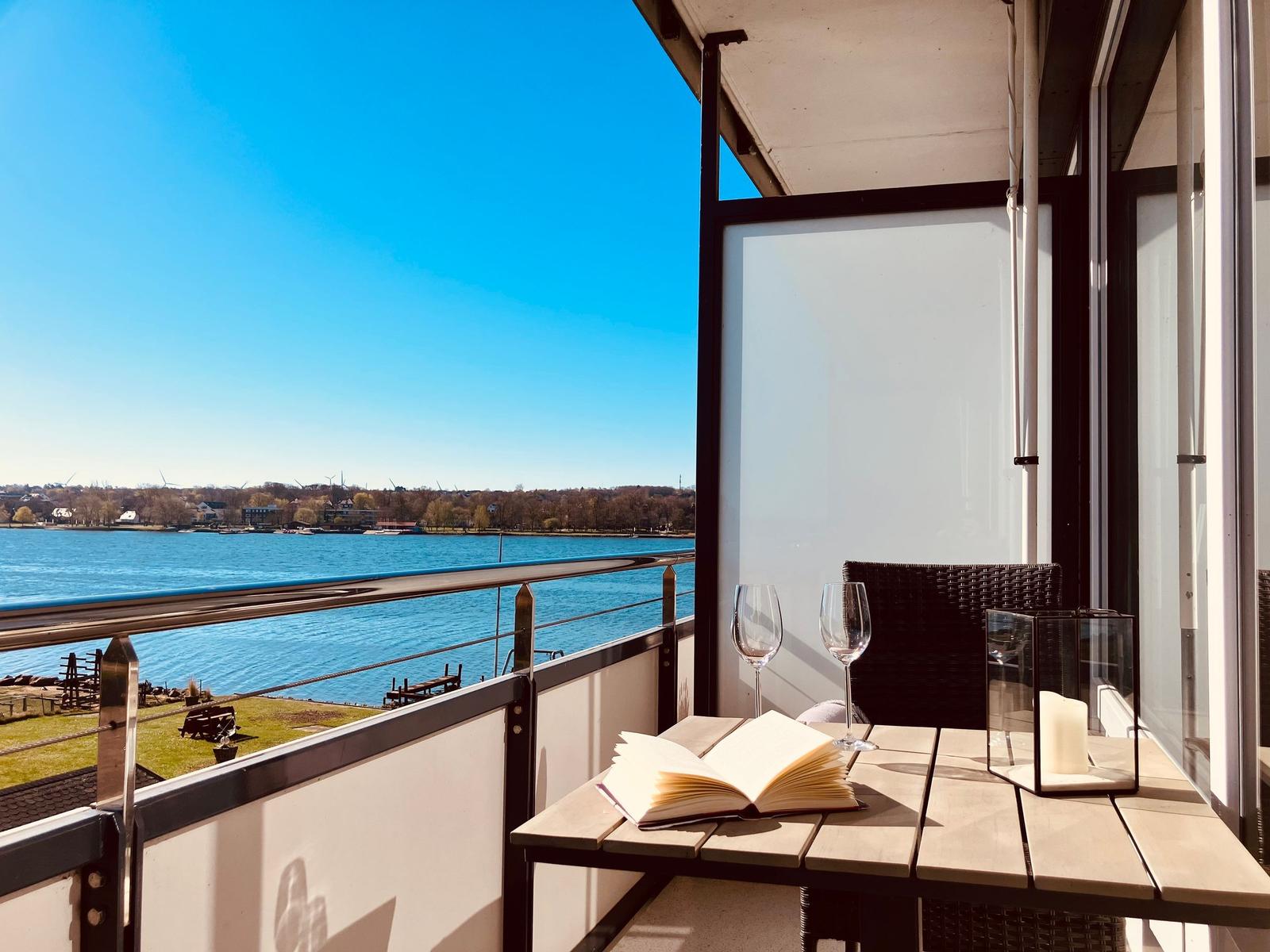 Balcony with table, chairs, and book overlooking a calm lake under clear blue sky.