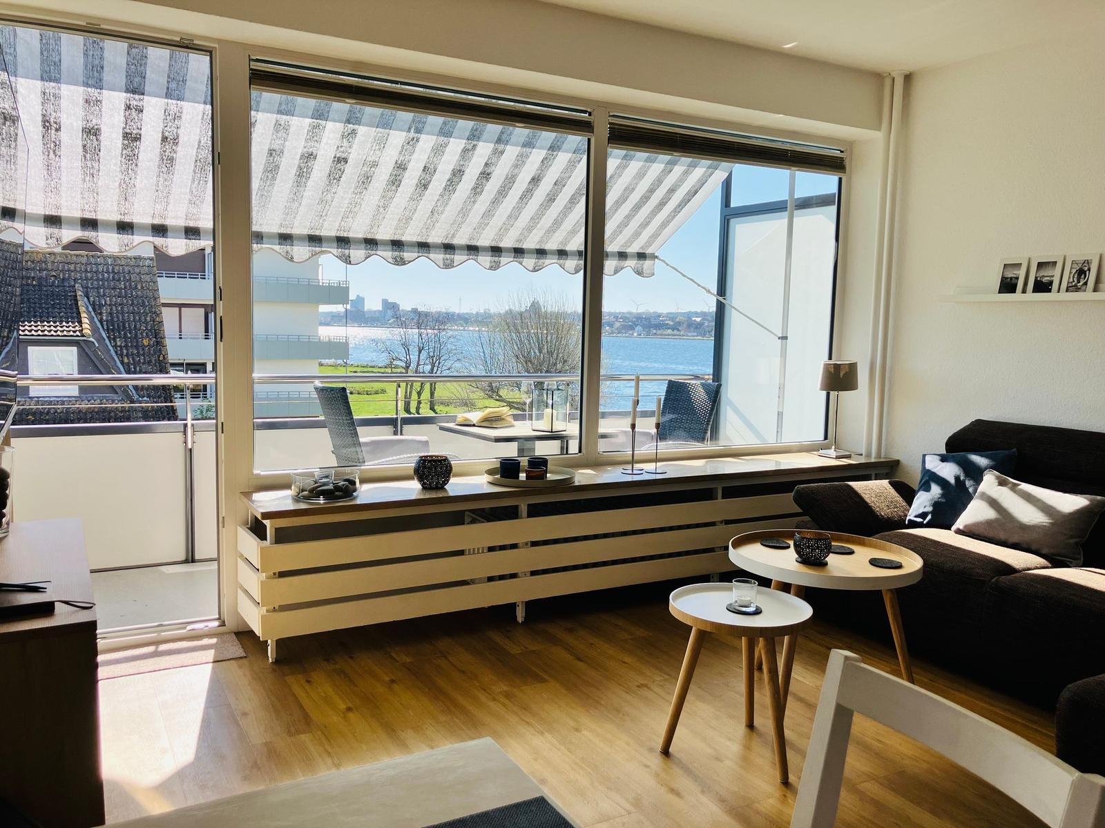 Wohnzimmer mit Balkon, Blick auf Wasser und Stadt. Holzboden, Sofa, Tische.