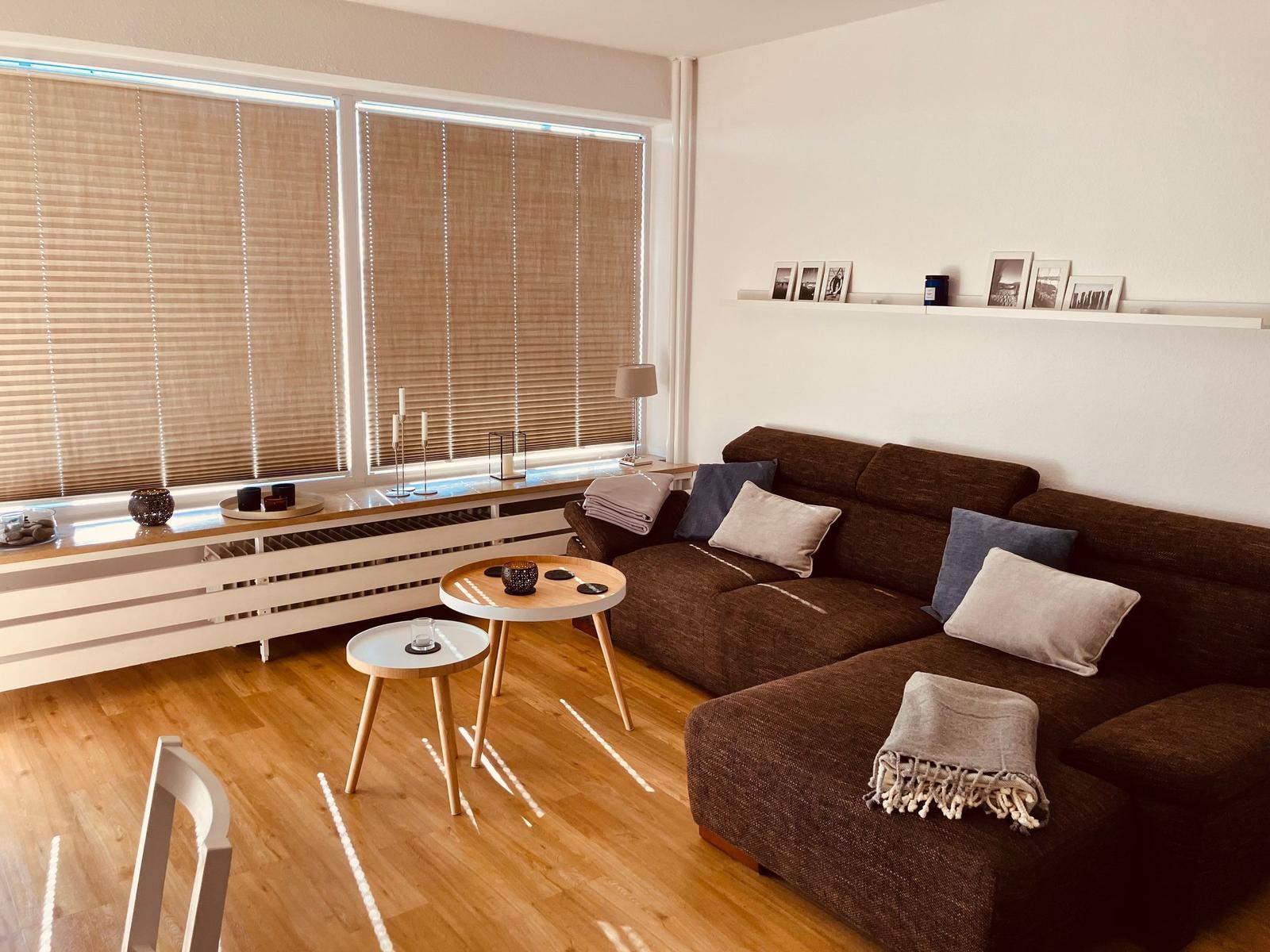 Ein Wohnzimmer mit braunem Sofa, zwei runden Tischen und Fenstern mit Jalousien.
