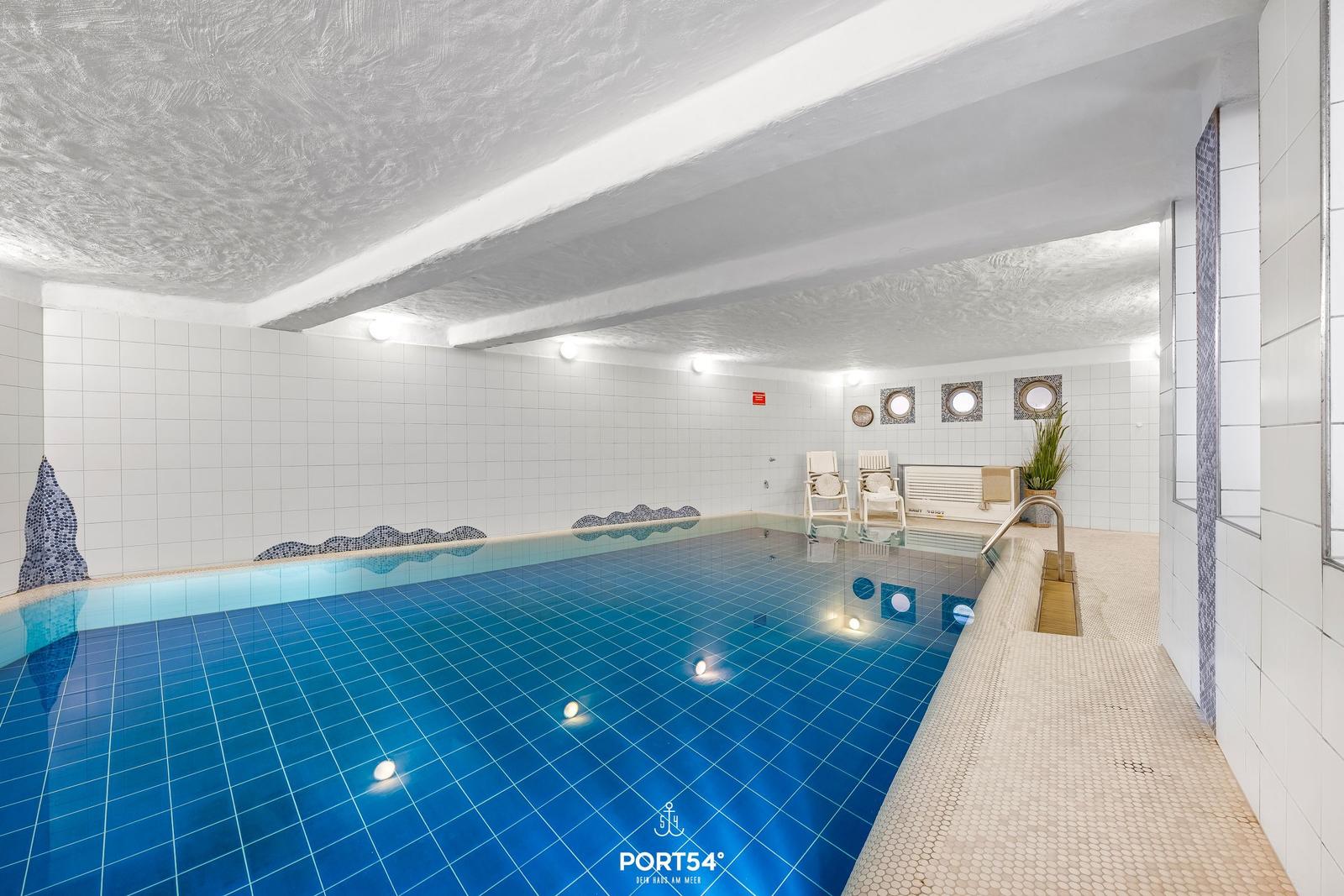 Indirekt beleuchteter Innenpool mit blauen Fliesen und weißen Wänden. Poolrand mit Liegestühlen.