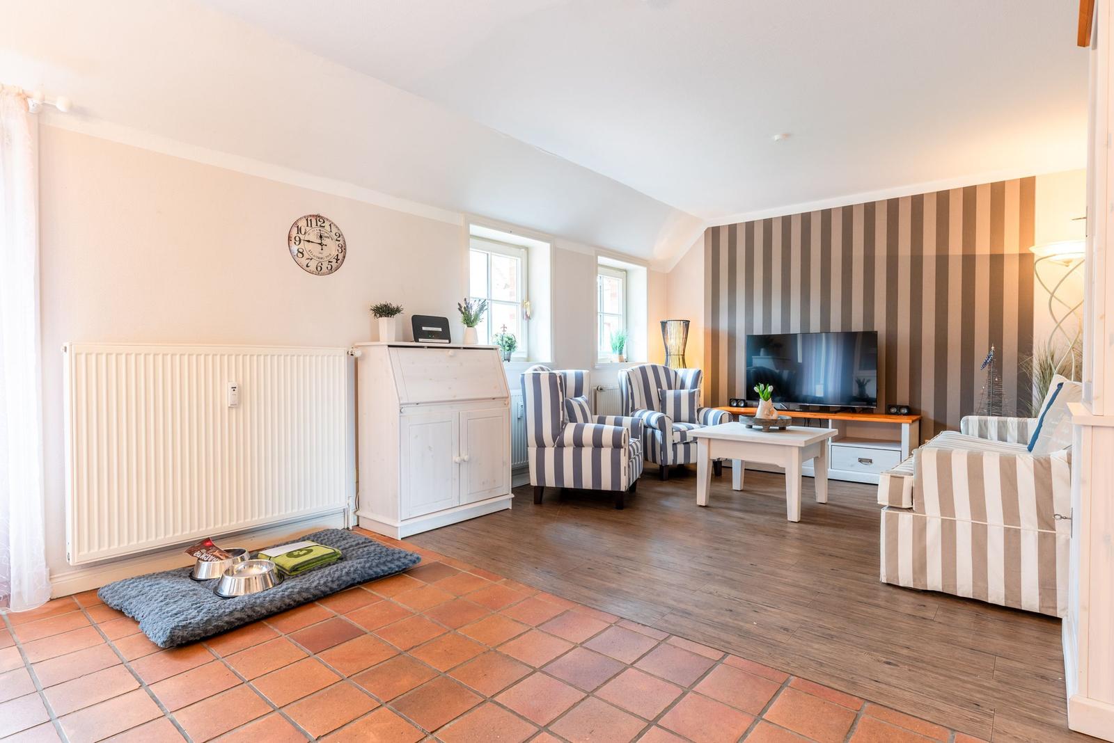 Wohnzimmer mit gestreiftem Sofa, Fernseher und Holzboden. Vorraum mit Terrakottafußboden und Hundebett.