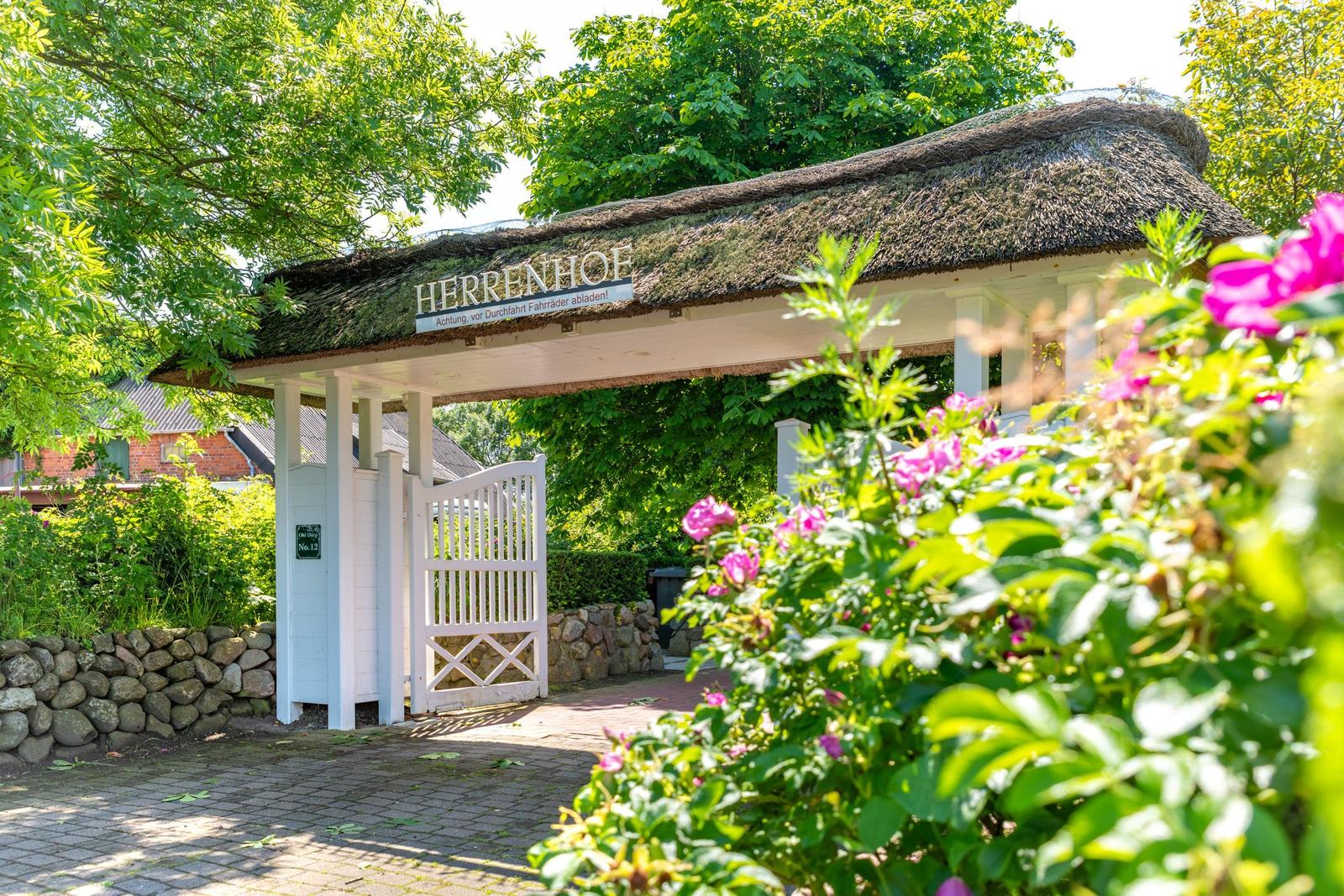 Eingangstor mit Strohdach und Schild Herrenhof. Rosenbusch im Vordergrund.