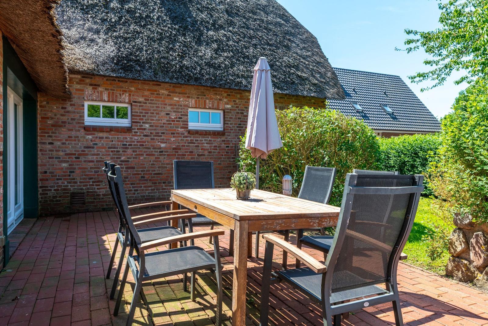 Terrasse mit Holztisch, Stühlen und geschlossenem Sonnenschirm vor einem strohgedeckten Haus.