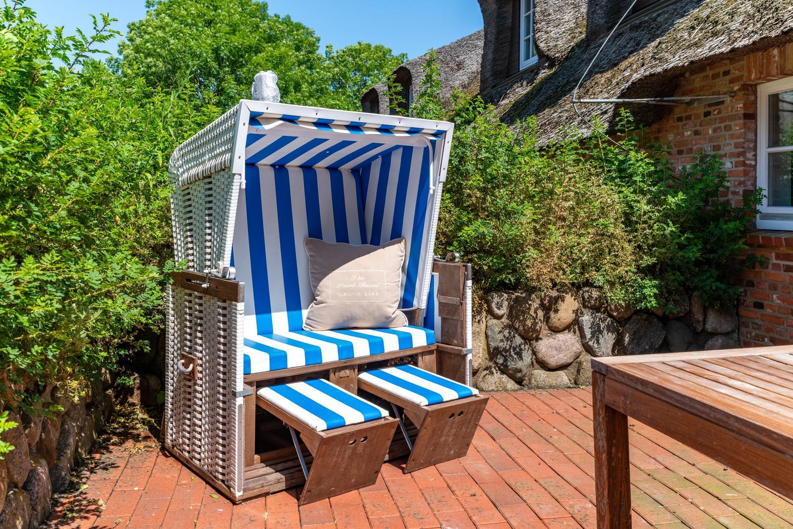 Blau-weiße Strandkörbe auf Terrasse vor traditionellem Haus mit Steinmauer und Grün.