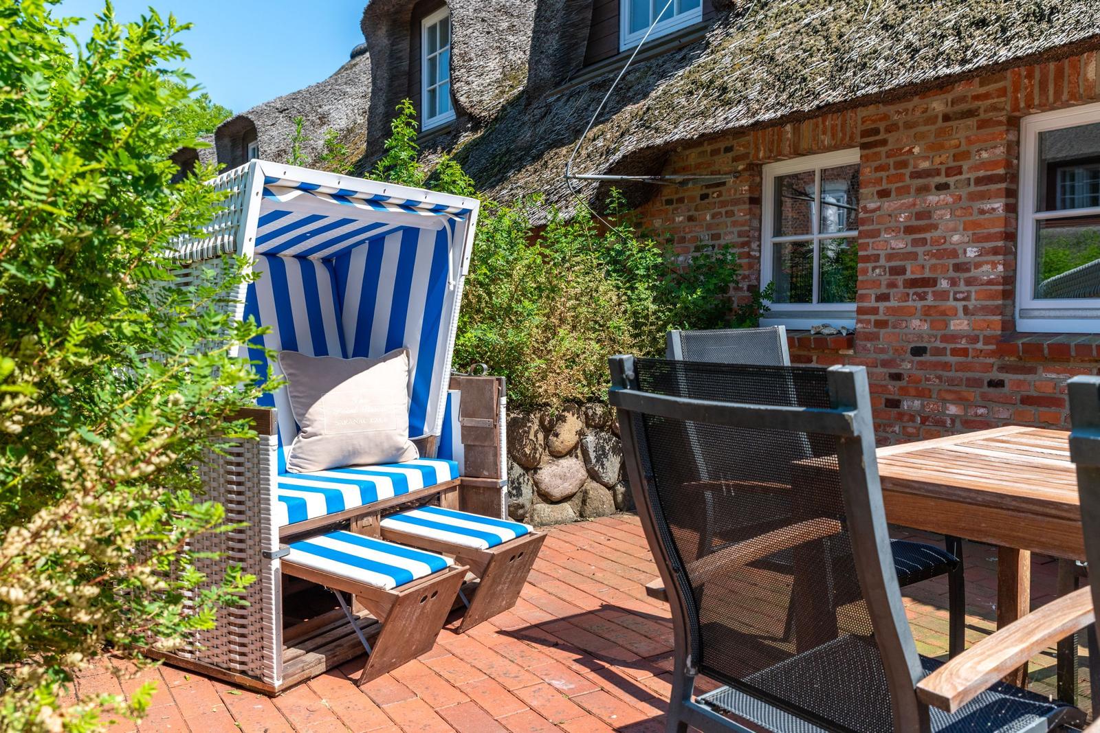 Terrasse mit Strandkorb, Tisch und Stühlen vor einem Steinhaus mit Dach aus Stroh.