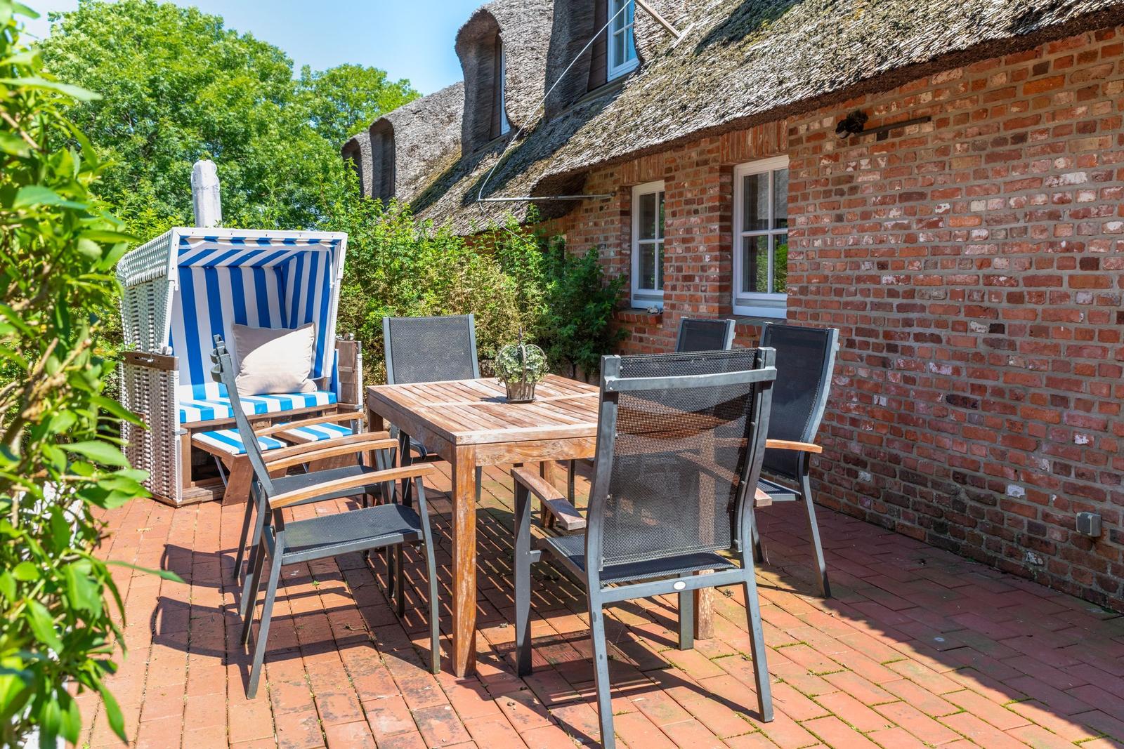 Terrasse mit Holztisch, Stühlen und Strandkorb vor einem Backsteinhaus mit Strohdach.