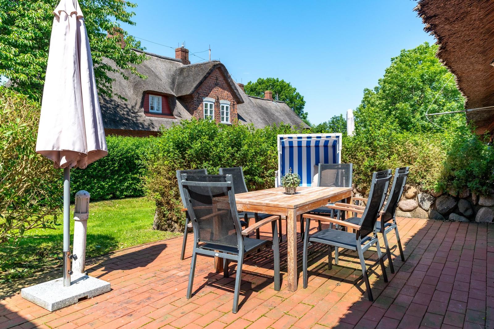 Terrasse mit Tisch, Stühlen und Sonnenschirm. Im Hintergrund ein Haus mit Strohdach.