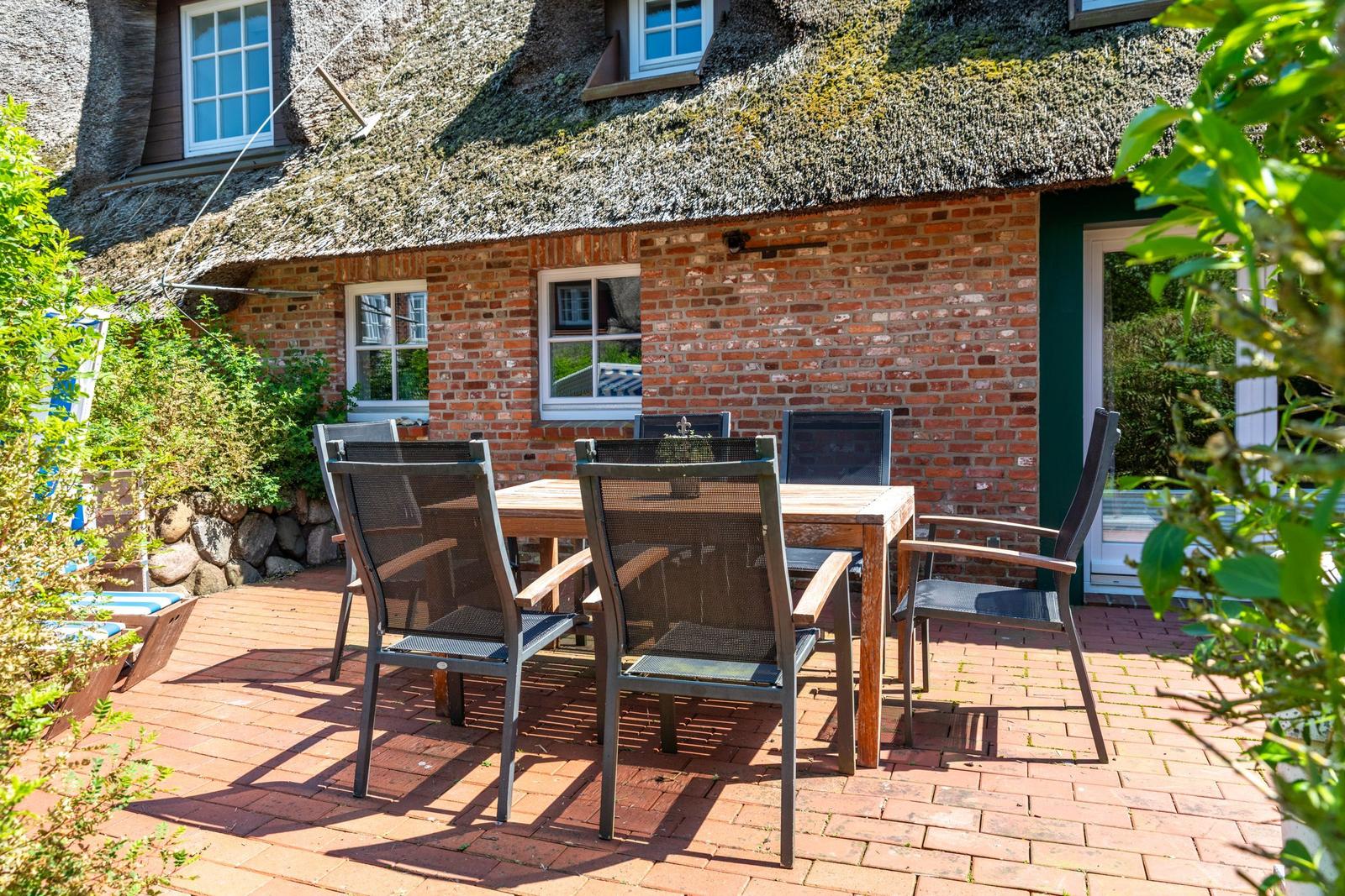 Terrasse mit Holztisch und Stühlen vor einem Haus mit strohgedecktem Dach und Backsteinmauer.