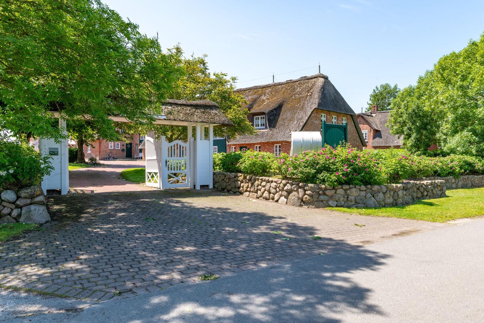 Haus mit Strohdach, weißer Toranlage und Blumenbeet hinter Steinmauer.