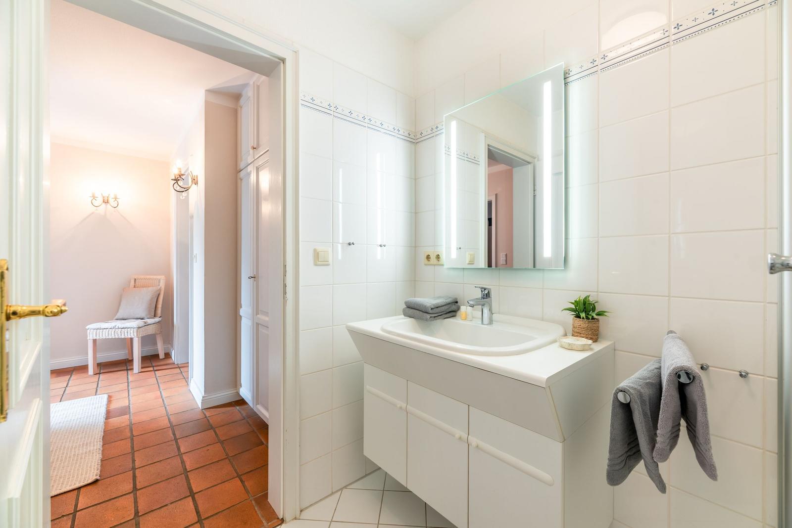 Badezimmer mit Waschbecken, Spiegel und Handtuchhalter. Durchgang zu einem Raum mit Stuhl.