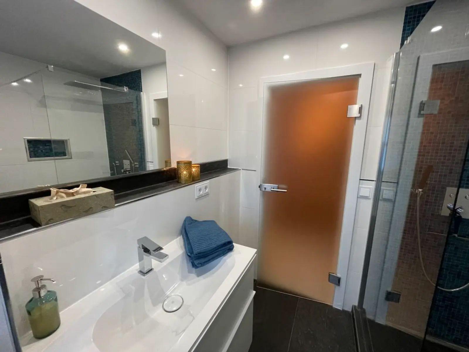 Badezimmer mit Waschbecken, Spiegel und Dusche. Tür führt in einen weiteren Raum.