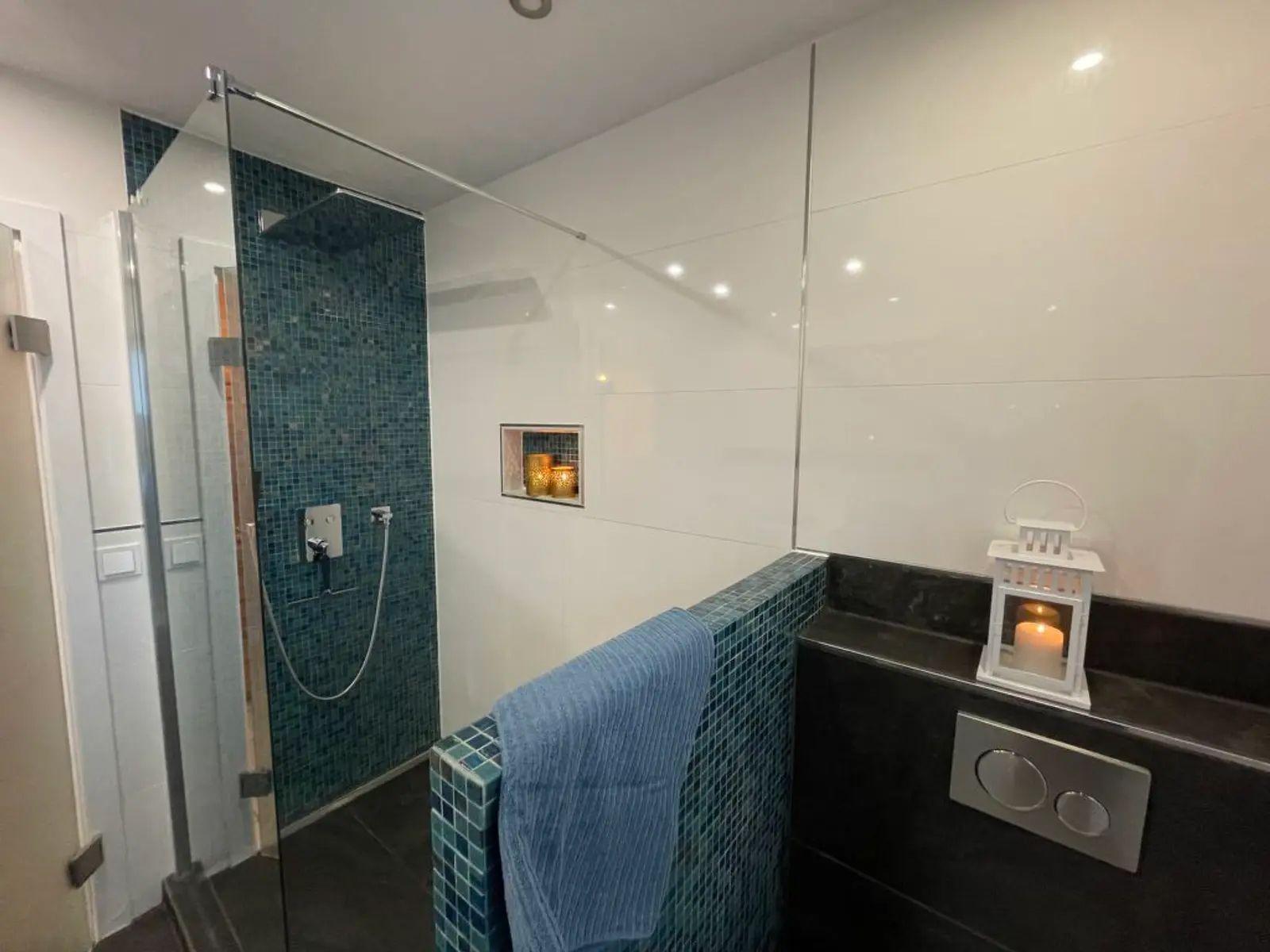 Dusche mit Glaswand und blauen Fliesen, daneben Toilette mit Kerze.