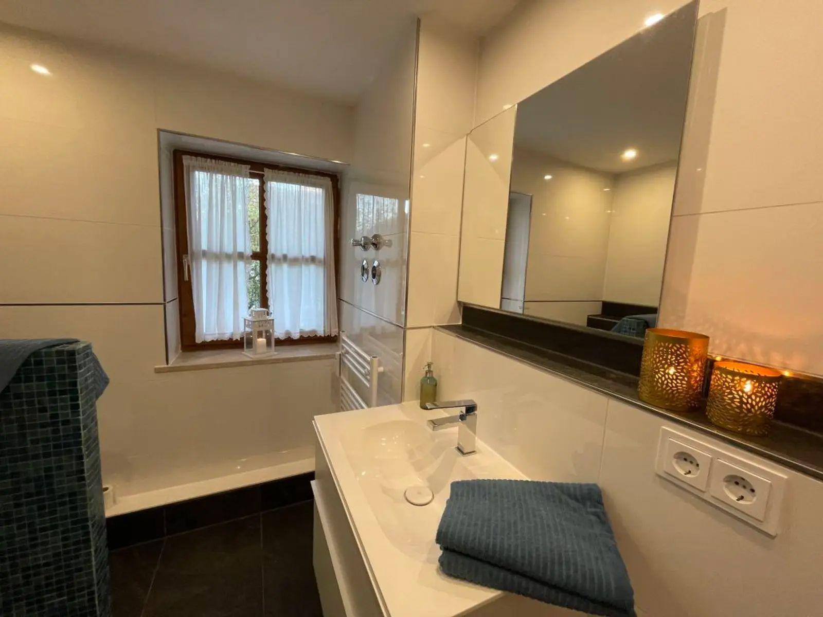 Badezimmer mit Waschbecken, Spiegel und Fenster. Auf der Arbeitsplatte stehen zwei Kerzen.