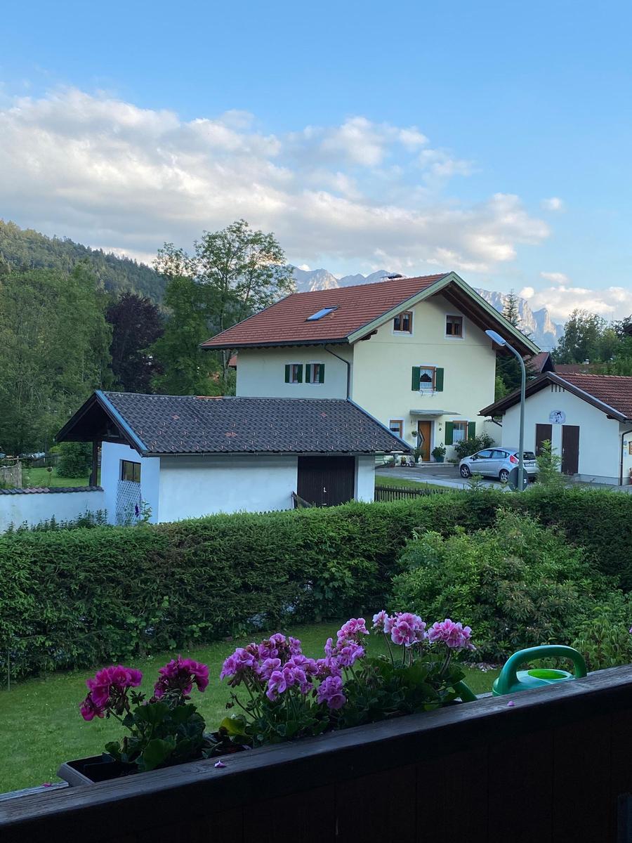 Haus mit rotem Dach, grüner Hecke und blauem Himmel mit Bergen im Hintergrund.