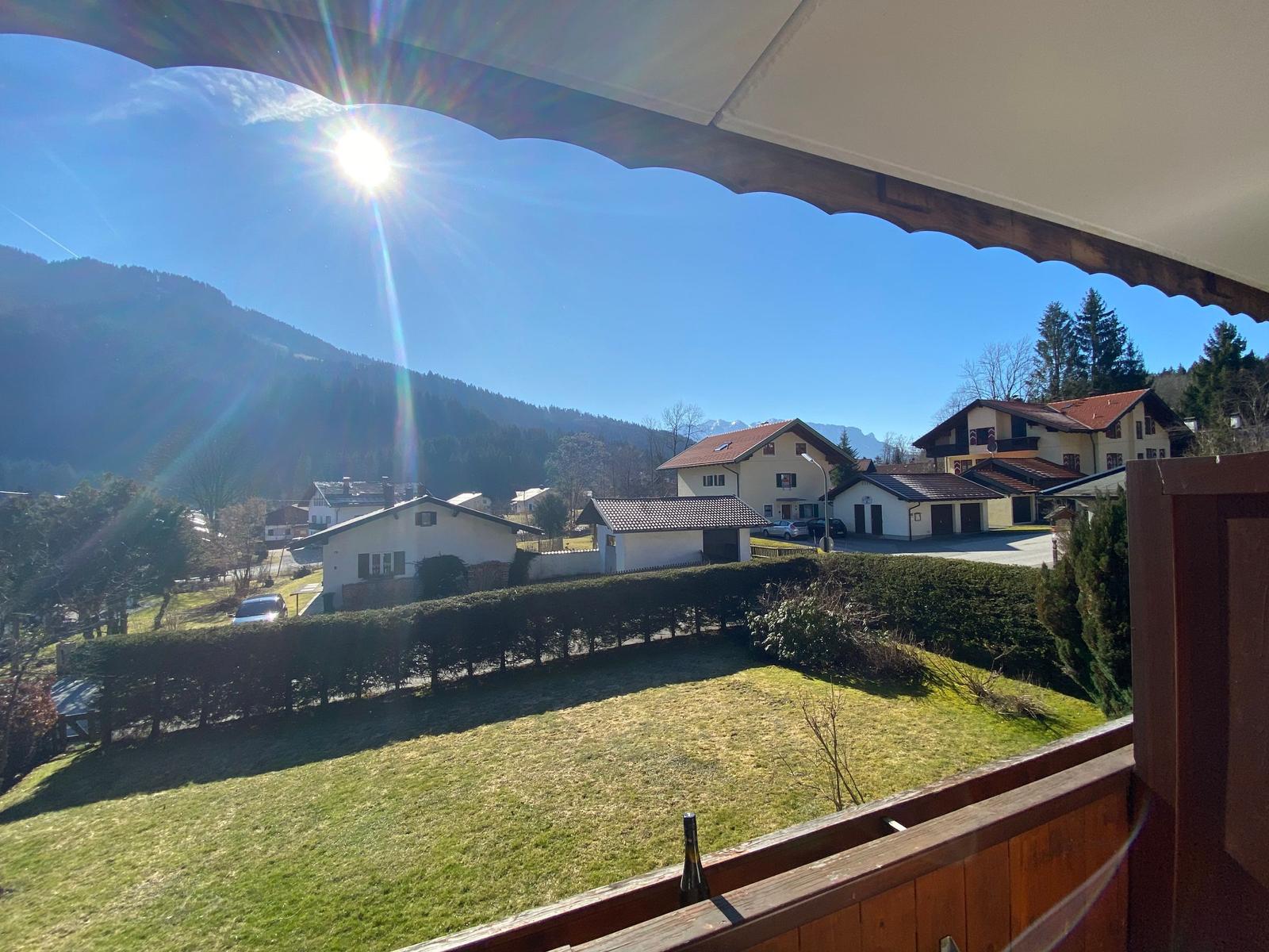 Blick von Balkon auf grünen Rasen, Hecken und Häuser mit Bergen im Hintergrund unter blauem Himmel.