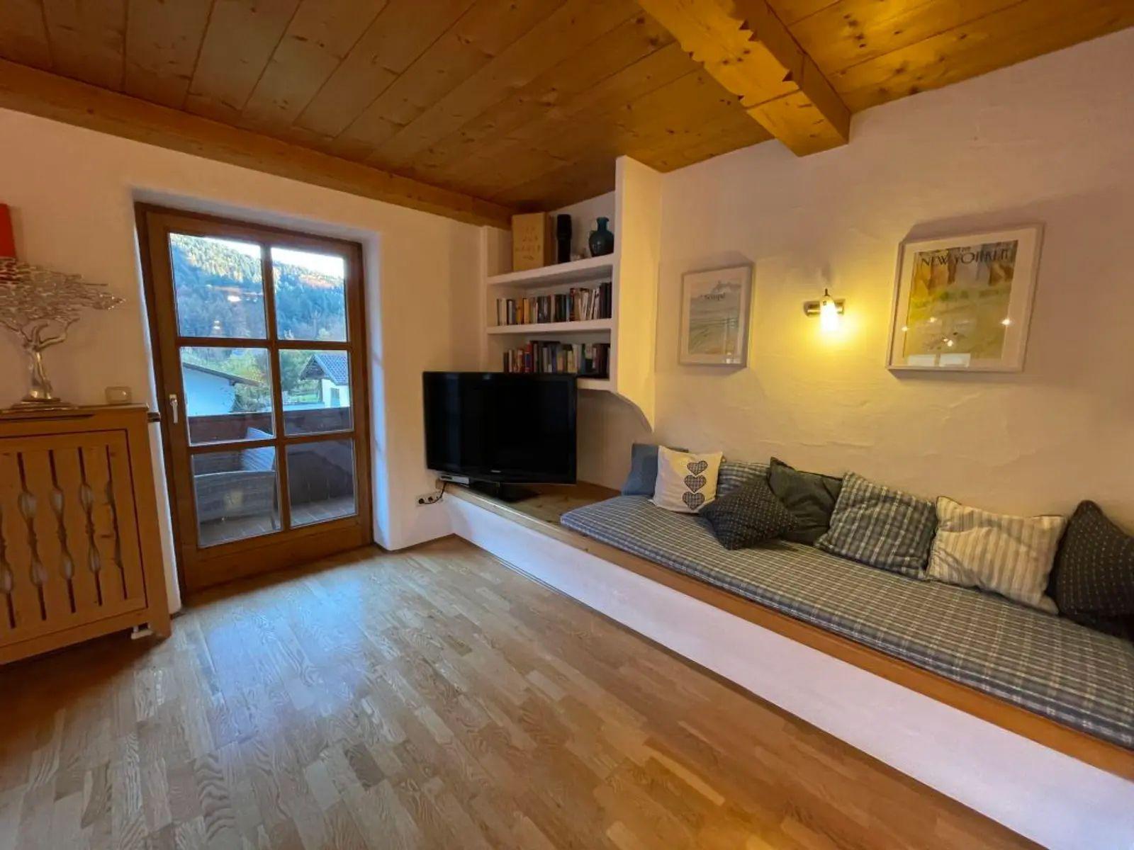 Wohnzimmer mit Sitzbank, Fernseher und Bücherregal. Holzdecke und Fenster zur Terrasse.