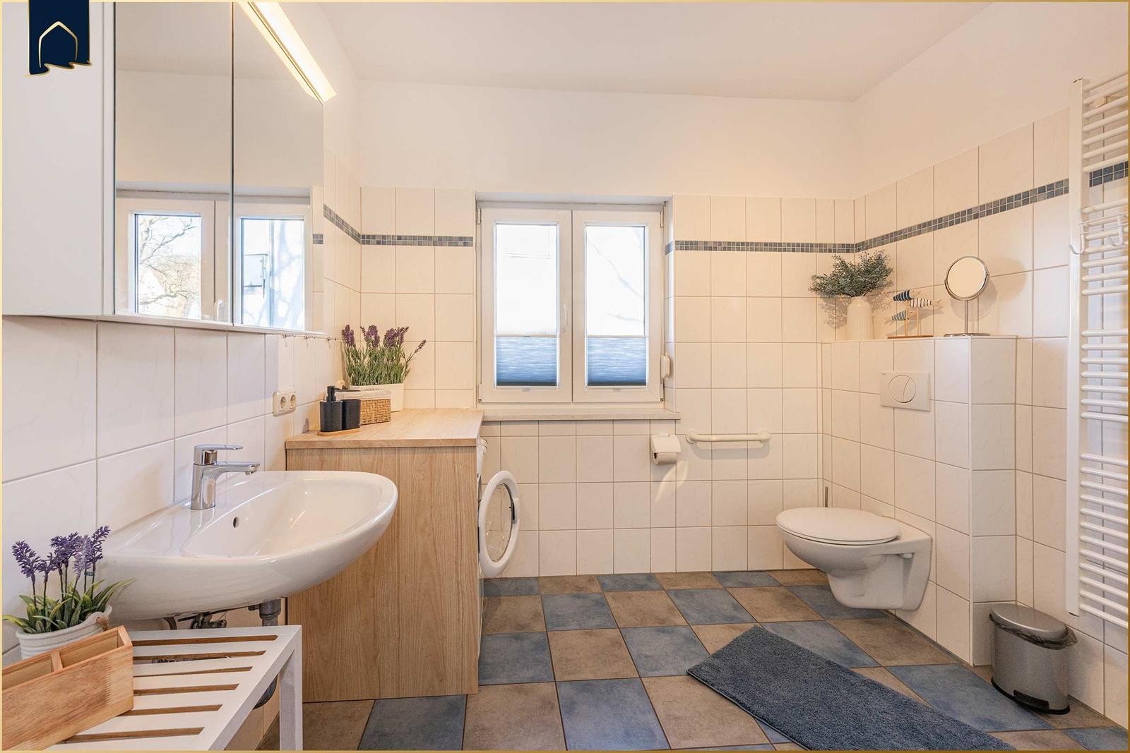 Badezimmer mit Waschbecken, Toilette und Fenster. Fliesenboden und Spiegel.