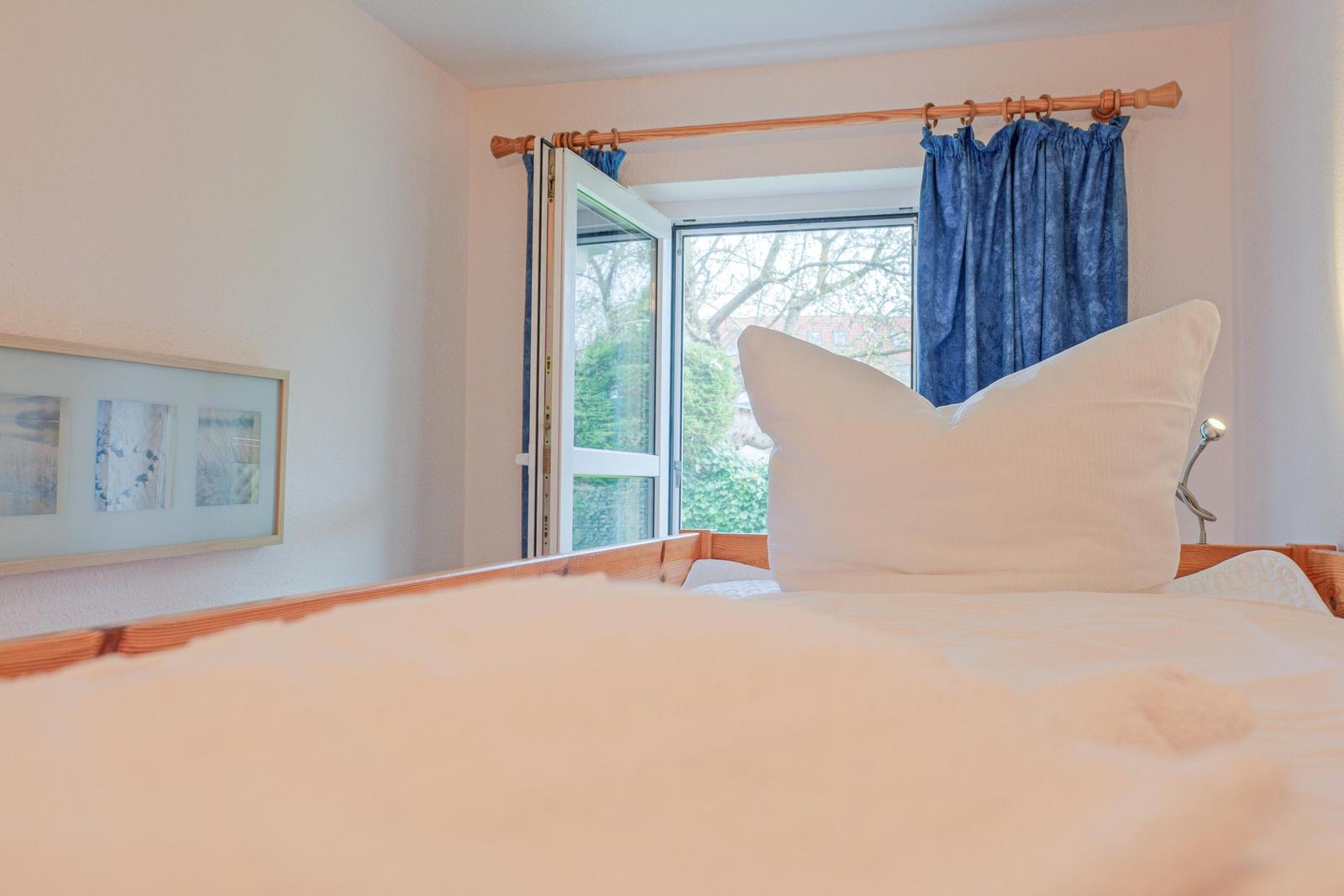 Ein Bett mit weißer Bettwäsche und Kissen. Ein Fenster mit blauen Vorhängen. Ein Bild an der Wand.