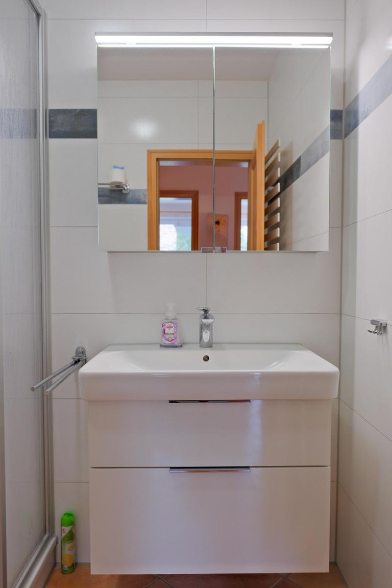 Bad mit Waschbecken, Spiegel und Dusche. Fliesenwand und Holztür sichtbar.