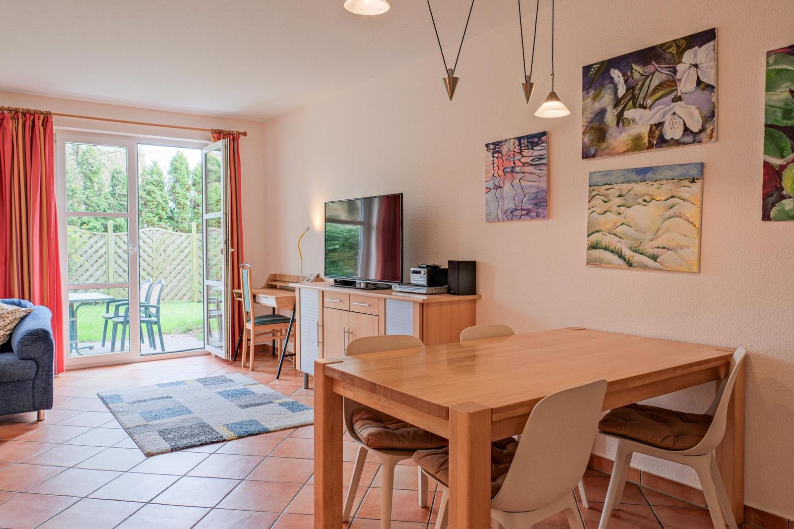 Ein Essbereich mit Holztisch und Stühlen. Im Hintergrund befindet sich ein Wohnbereich mit TV und Fenster.