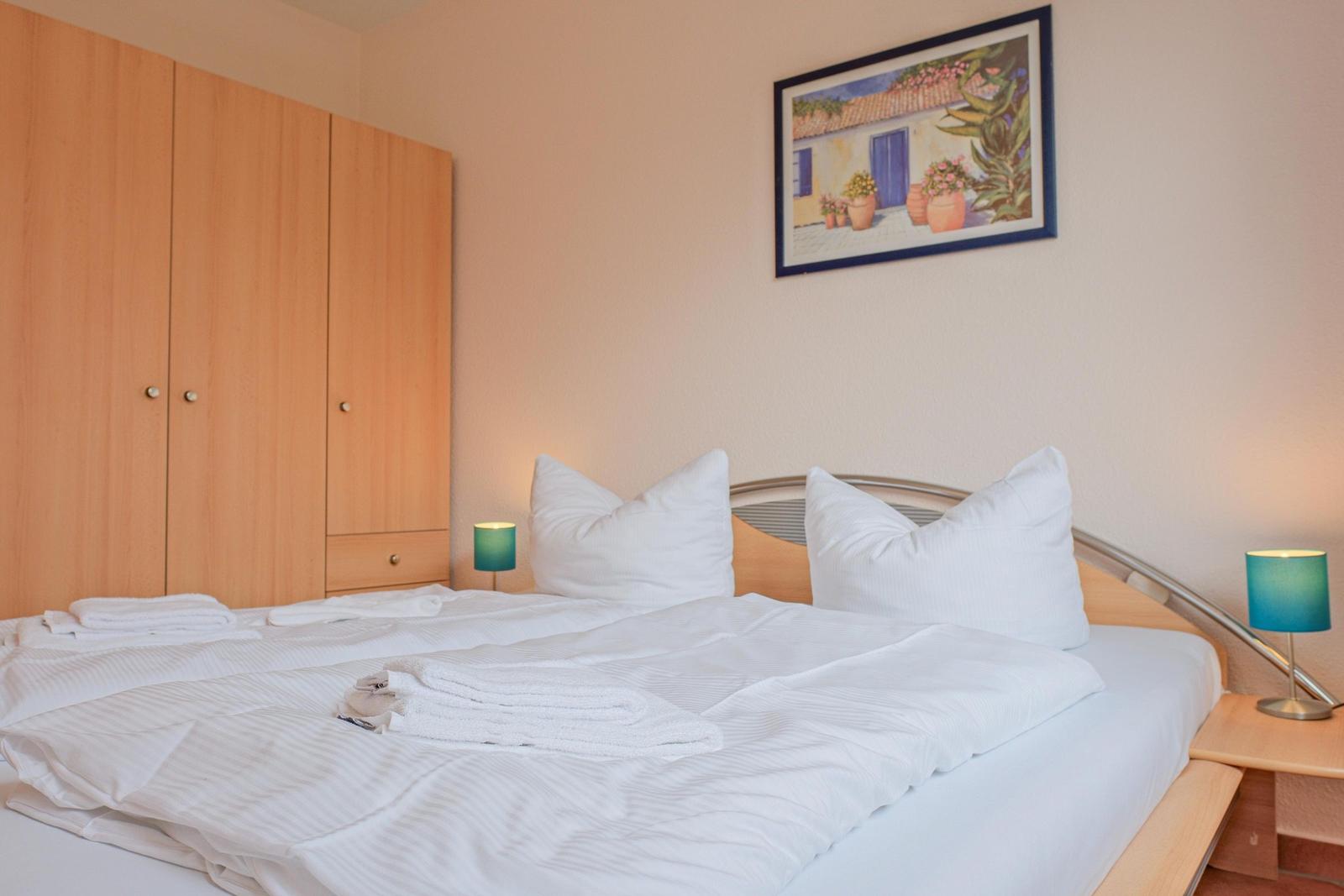 Ein Bett mit weißen Laken und Kissen. An der Wand hängt ein Bild. Links steht ein Schrank.