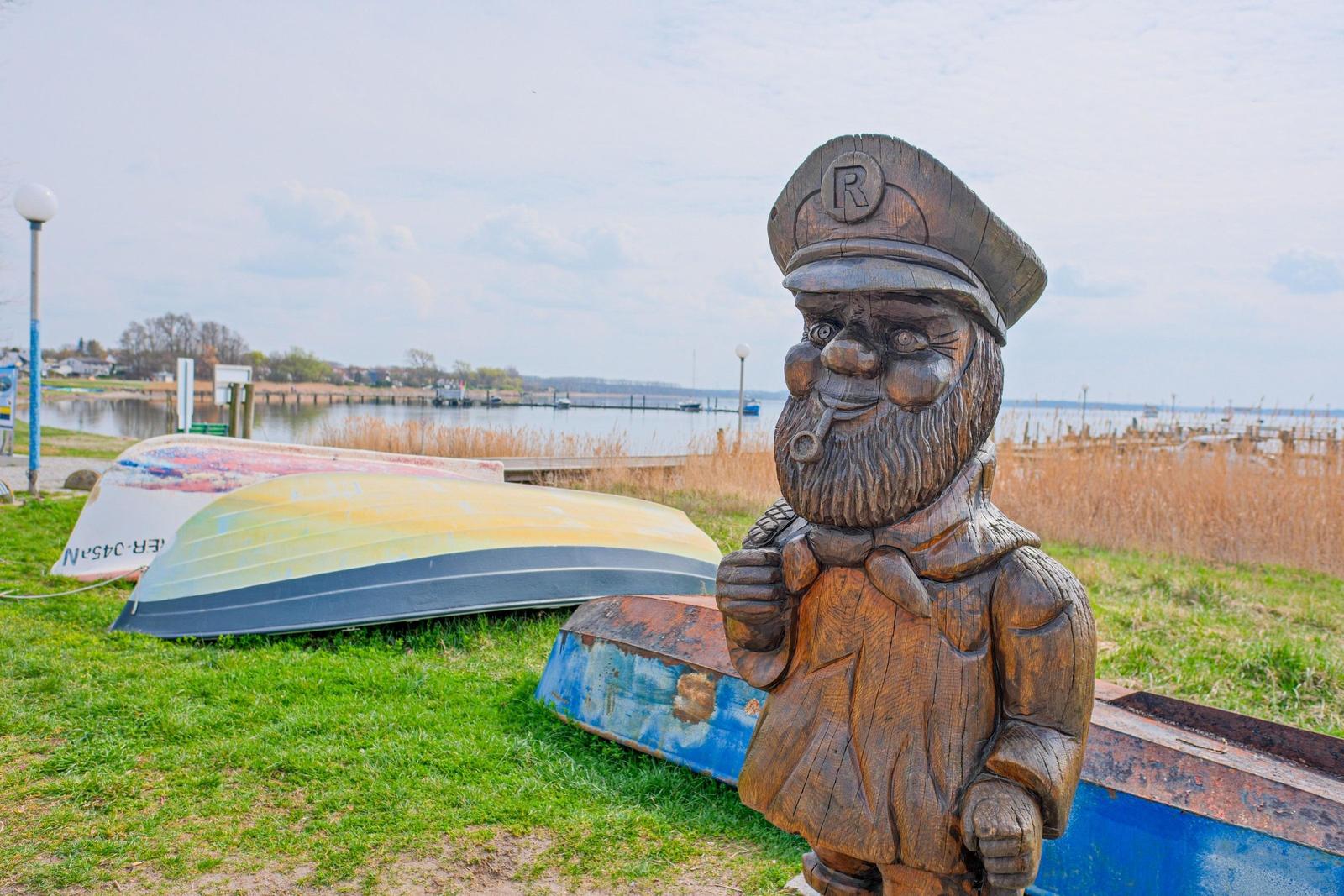 Holzfigur eines Steuermanns mit Pfeife neben Booten am Ufer.