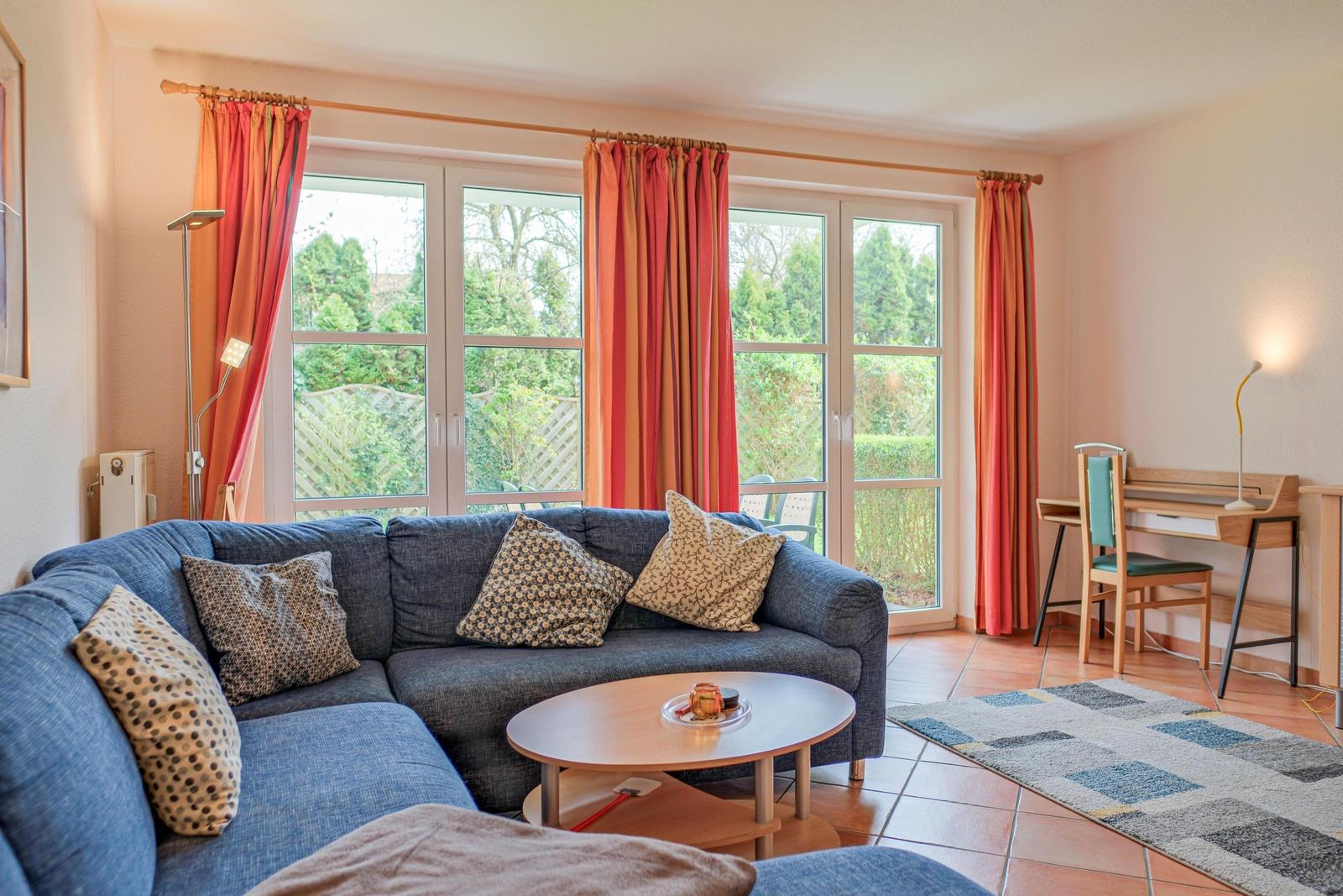 Ein Wohnzimmer mit Sofa, Tisch und Schreibtisch. Große Fenster mit orangefarbenen Vorhängen.
