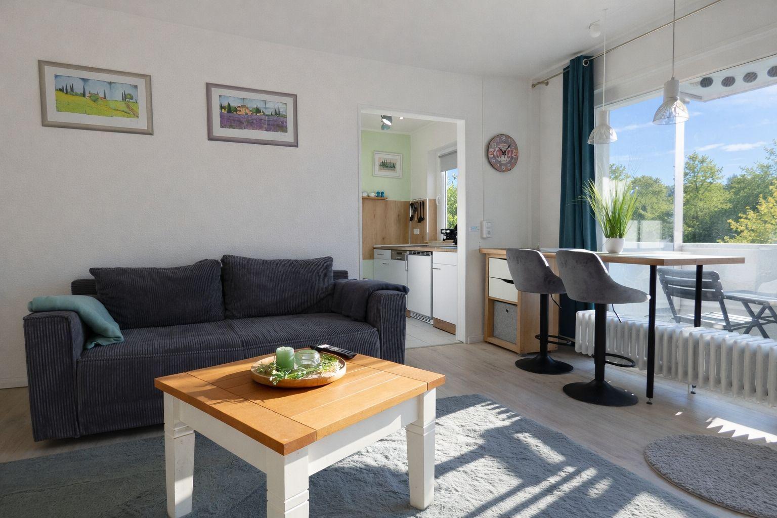Wohnbereich mit Sofa, Tisch und Blick auf Balkon. Küche im Hintergrund.