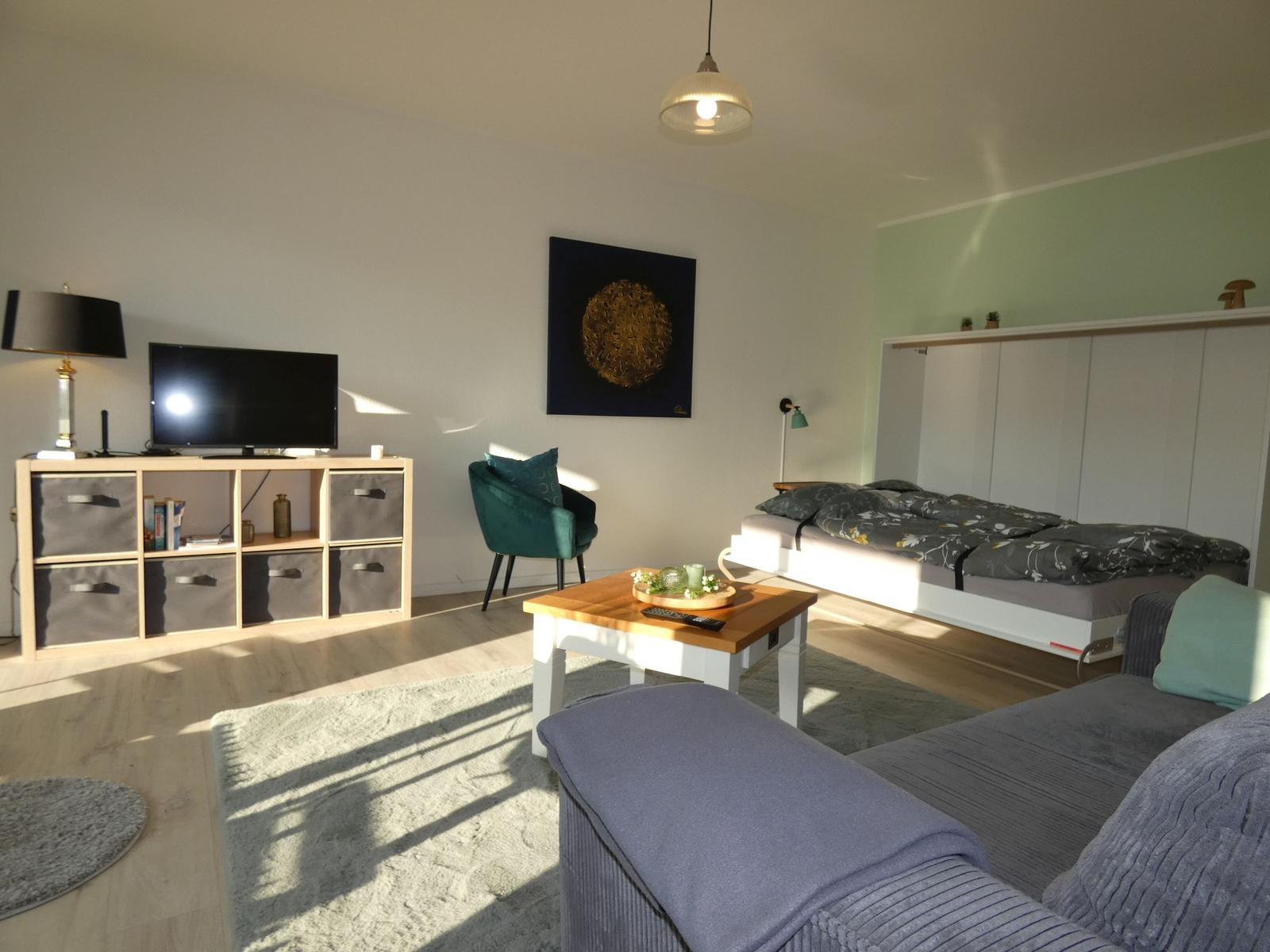 Ein Raum mit Sofa, Bett, TV und Tisch. Wandbild und grüne Wand sind sichtbar.