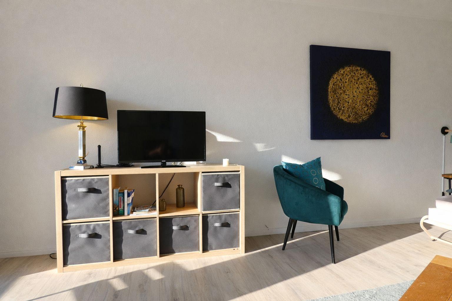 Ein Wohnzimmer mit einem Fernseher auf einem Holzregal, einem blauen Sessel und einer Wanddekoration.