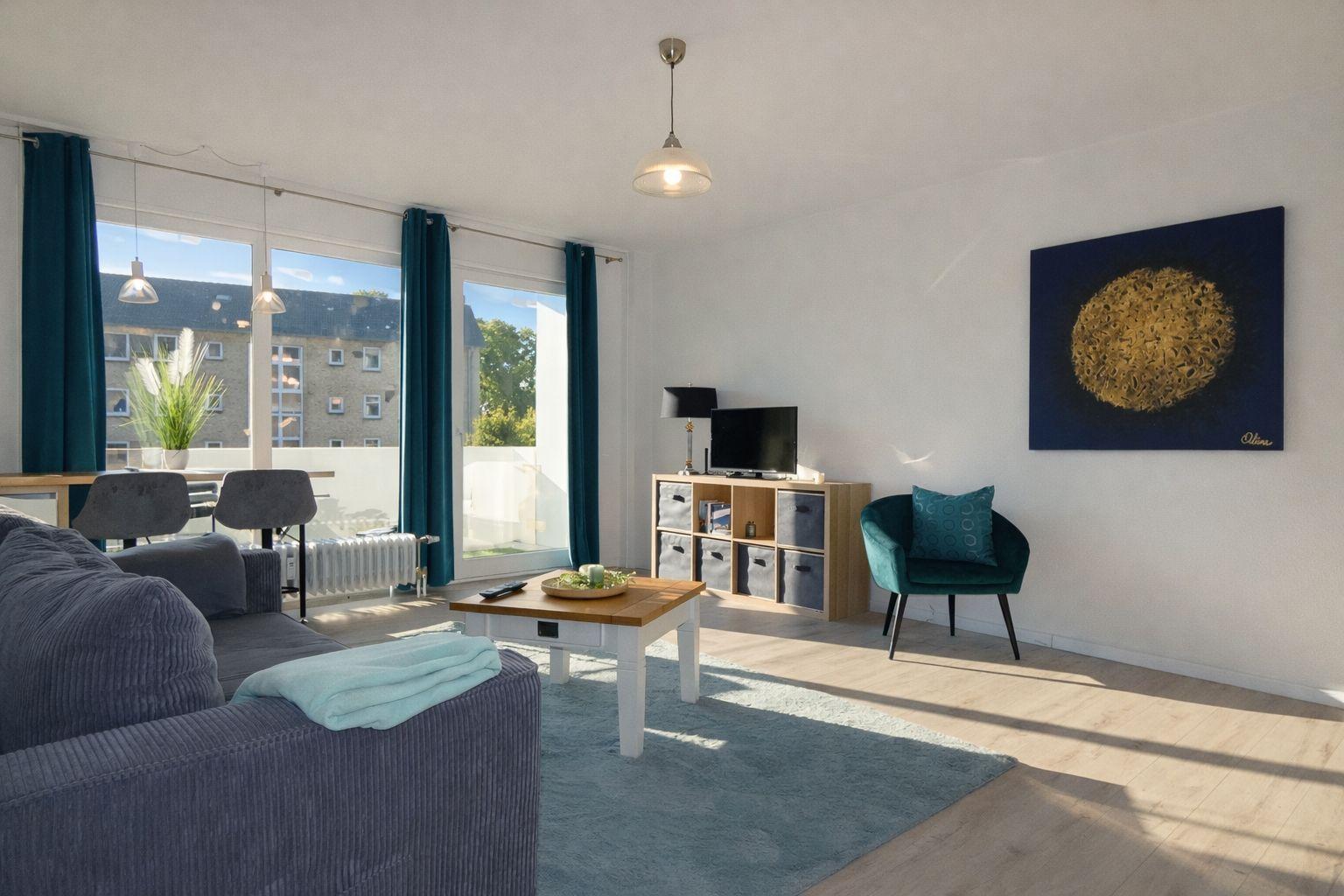 Wohnzimmer mit Sofa, Sessel, Tisch und TV. Große Fenster mit Blick auf Balkon.