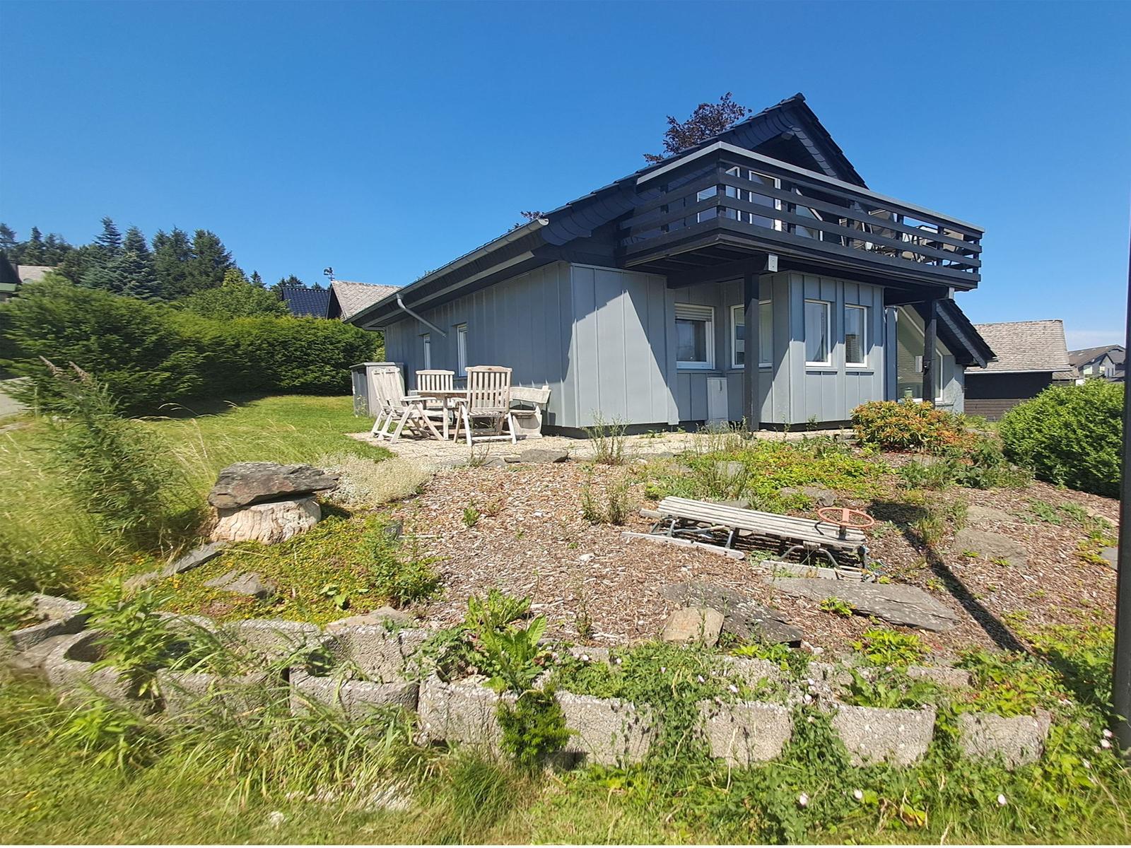 Ein Haus mit Balkon, Garten und Holzmöbel unter blauem Himmel.