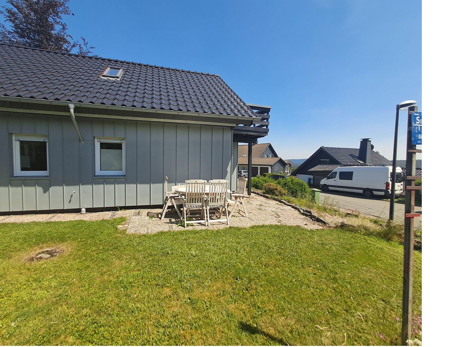Haus mit Terrasse, Garten und Tisch mit Stühlen. Hintergrund: Straße mit Wohnhäusern.