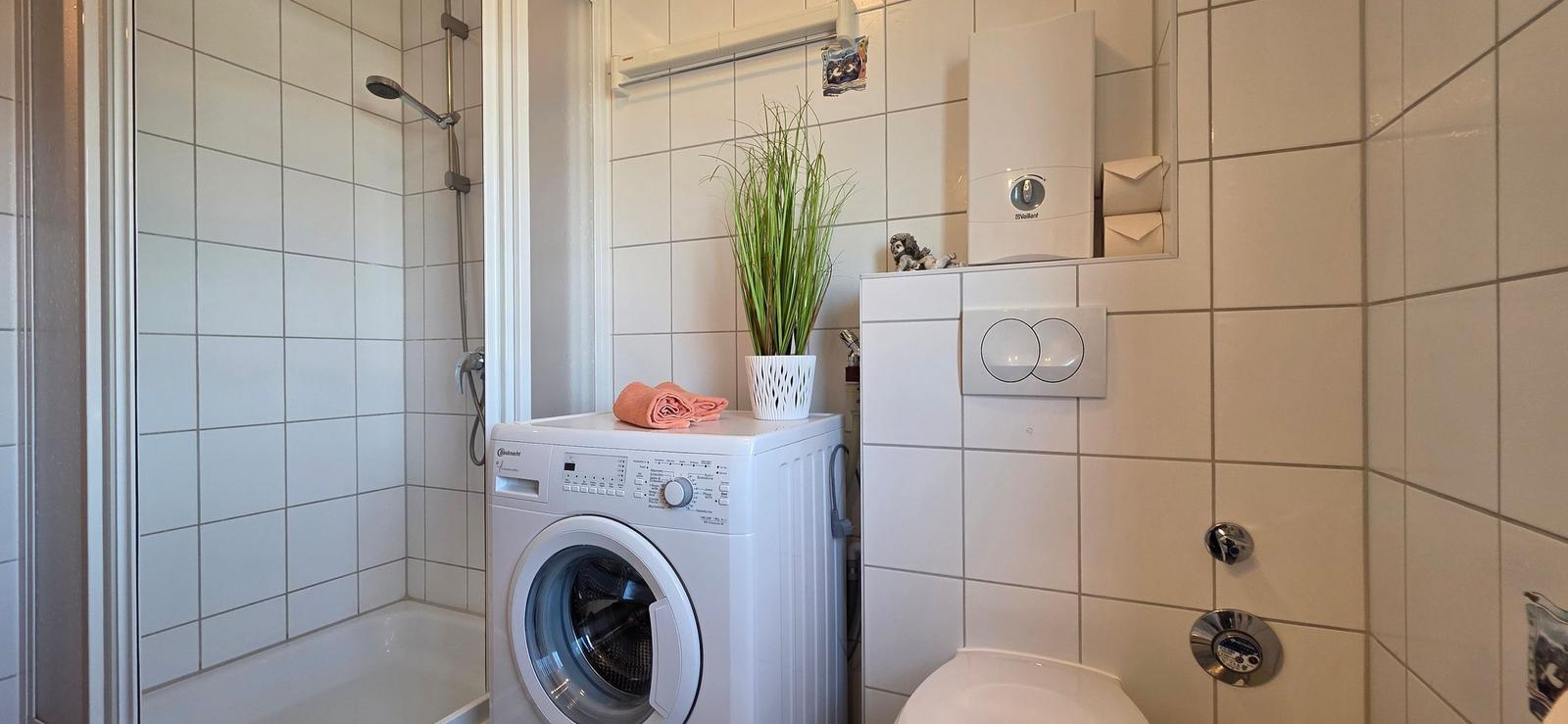 Badezimmer mit Dusche, Waschmaschine und Toilette. Eine Pflanze steht auf der Maschine.