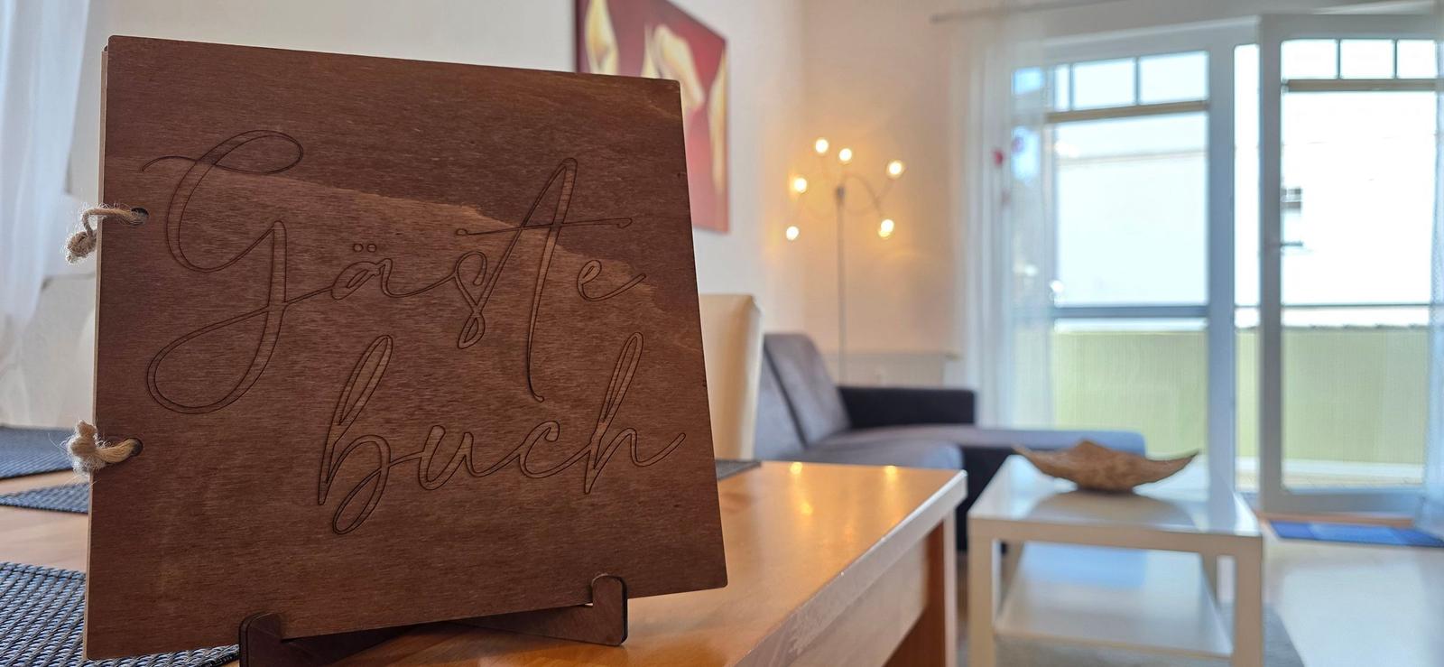 Holz-Gästebuch auf Tisch, dahinter Wohnbereich mit Sofa und Fenster.