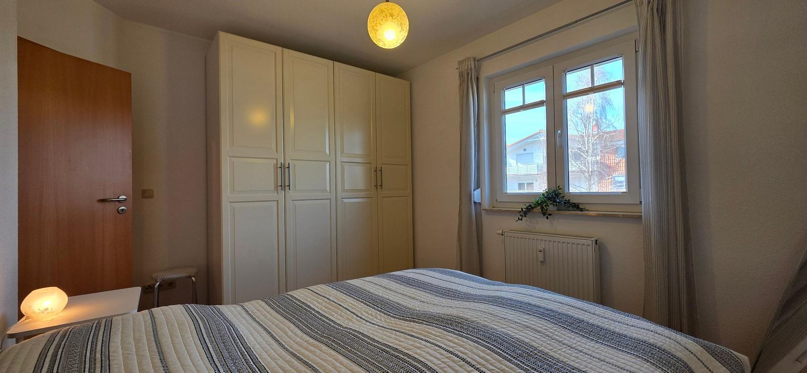 Schlafzimmer mit Bett, Kleiderschrank und Fenster mit Blick auf Außenbereich.