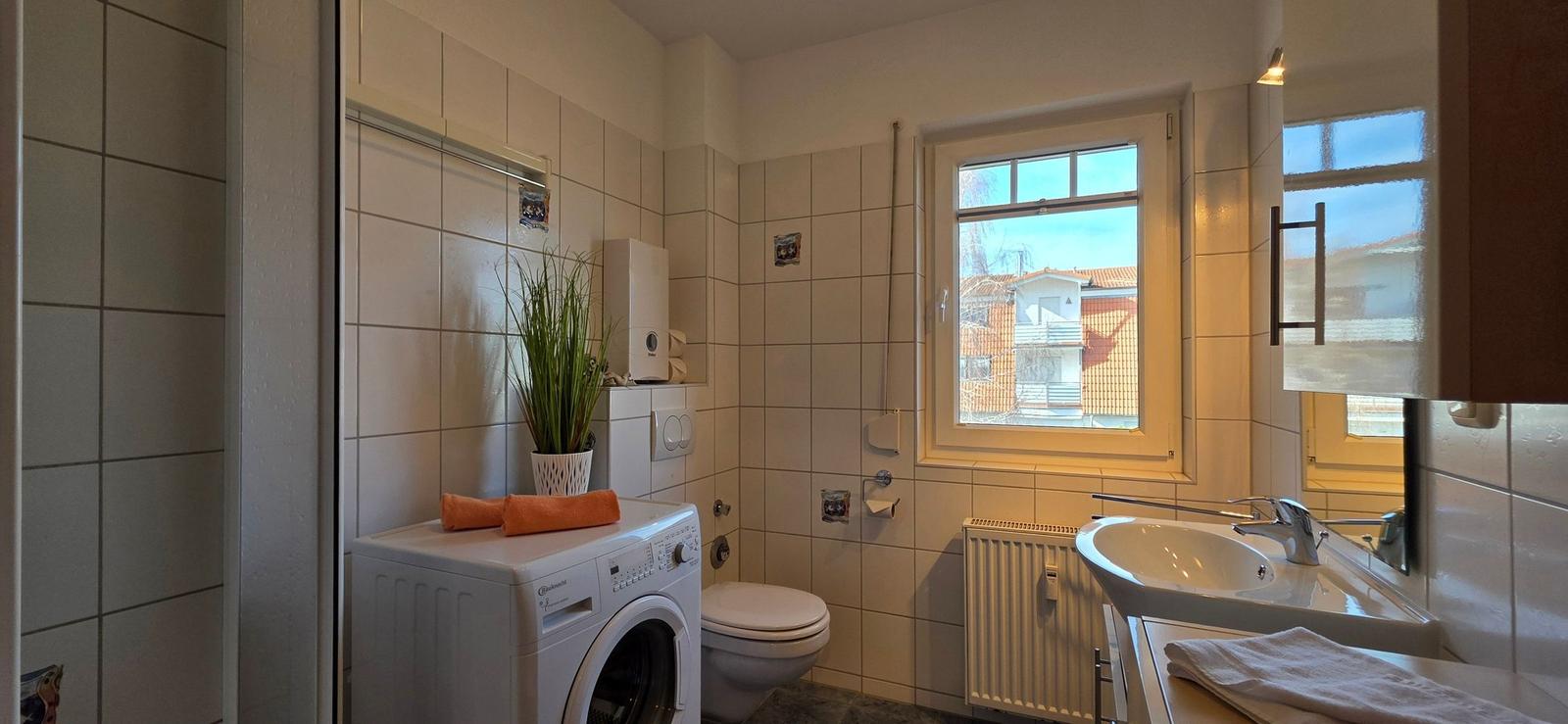 Badezimmer mit Waschmaschine, Toilette und Waschbecken. Fenster mit Blick auf Haus.