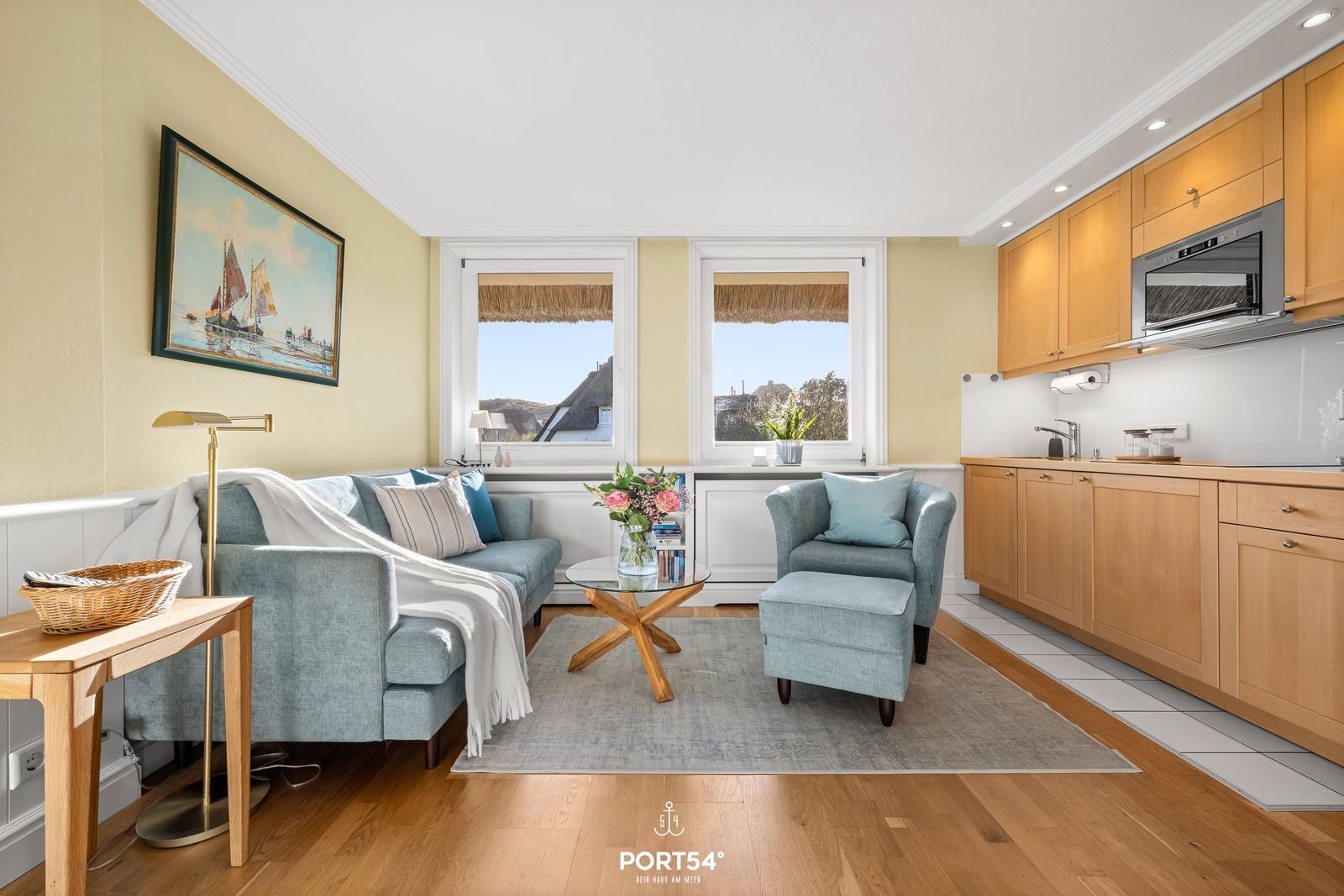 Wohnküche mit Sofa, Sessel und Küchenzeile. Fenster mit Blick auf Dächer.